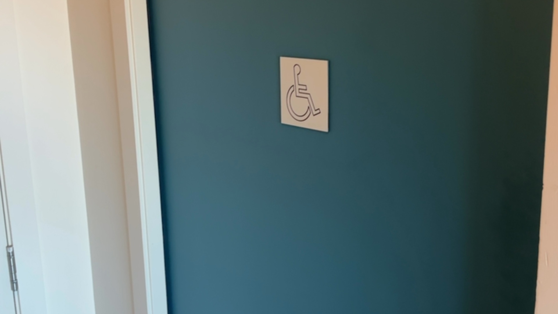 Teal-coloured door with a white square sign featuring a dark outline of a wheelchair symbol, indicating an accessible facility.