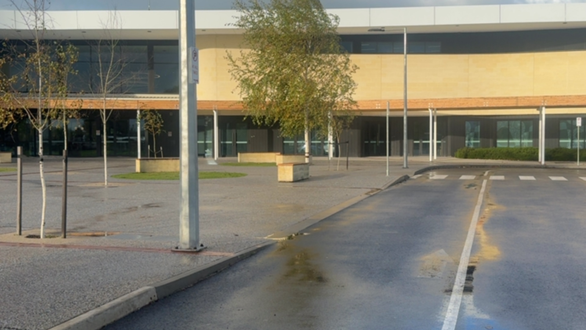 Level asphalt and concrete forecourt leading to a tan building with glass entrances. Includes a marked zebra crossing and flat pedestrian area with block-style concrete benches.