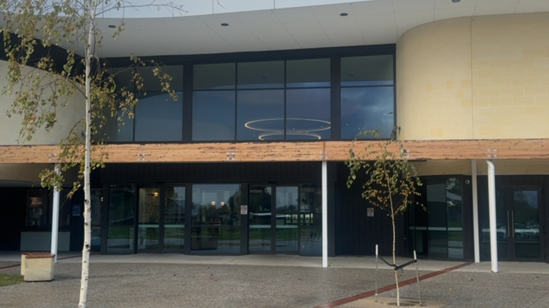 Level paved forecourt leads to automatic sliding glass doors at grade. Wide wooden beam above entry supported by white pillars. A concrete bench is located on the left.