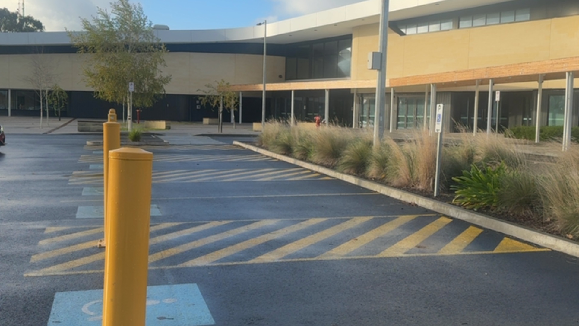 Accessible parking with blue wheelchair symbols, yellow bollards and hatched markings. Level asphalt surface and concrete kerb lead to a modern building with a covered walkway entrance.