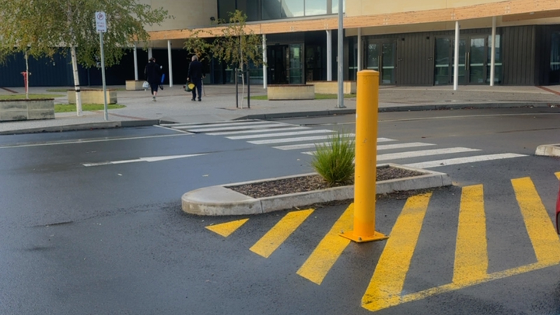 White zebra crossing on wet asphalt leads to a flat concrete ramp and paved area. A tall yellow bollard stands on yellow hatched road markings near the level building entrance.