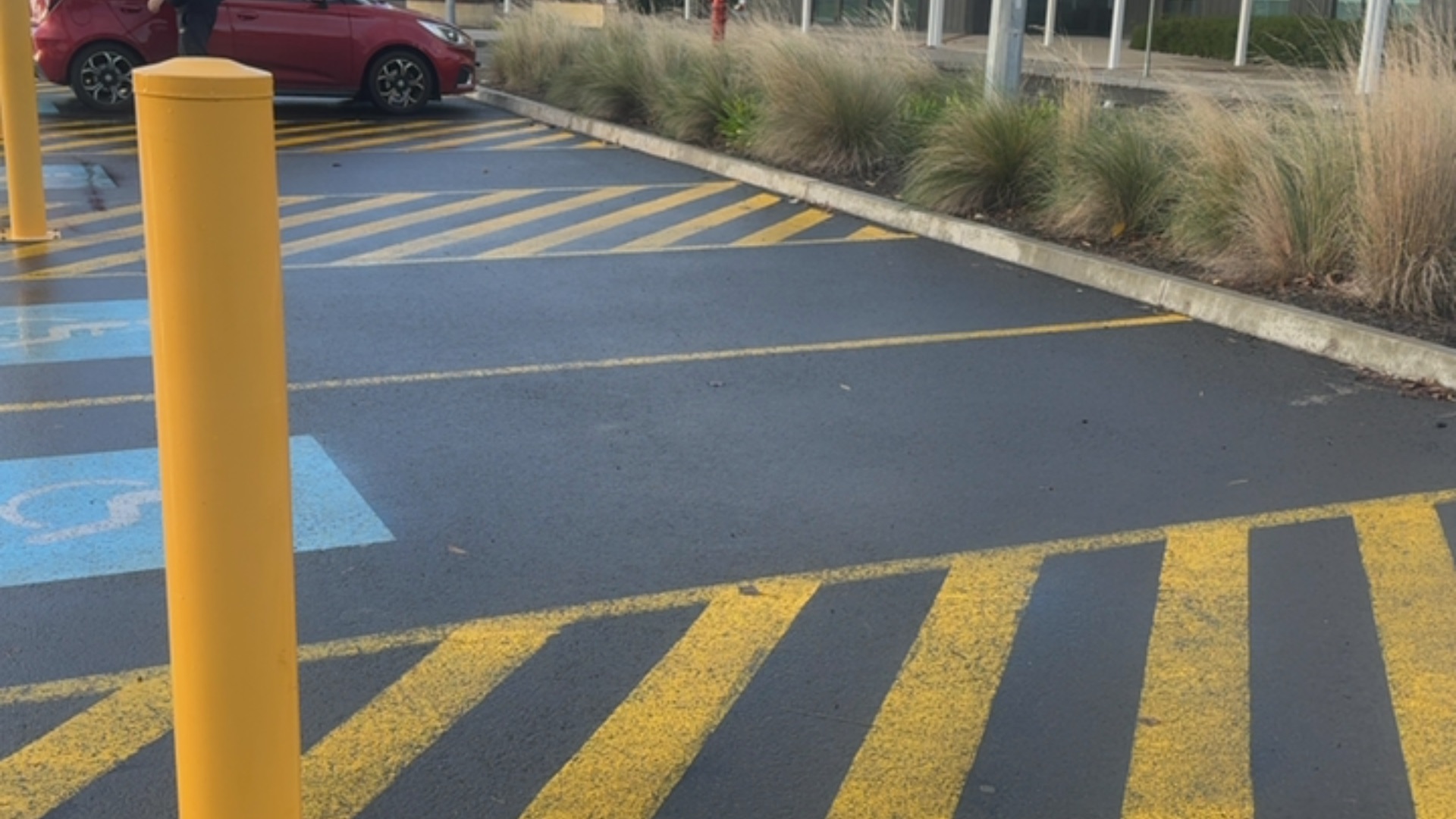 Level asphalt parking area with yellow hatched markings and blue wheelchair symbol. A tall yellow bollard is in the foreground next to a concrete kerb and landscaped garden bed.