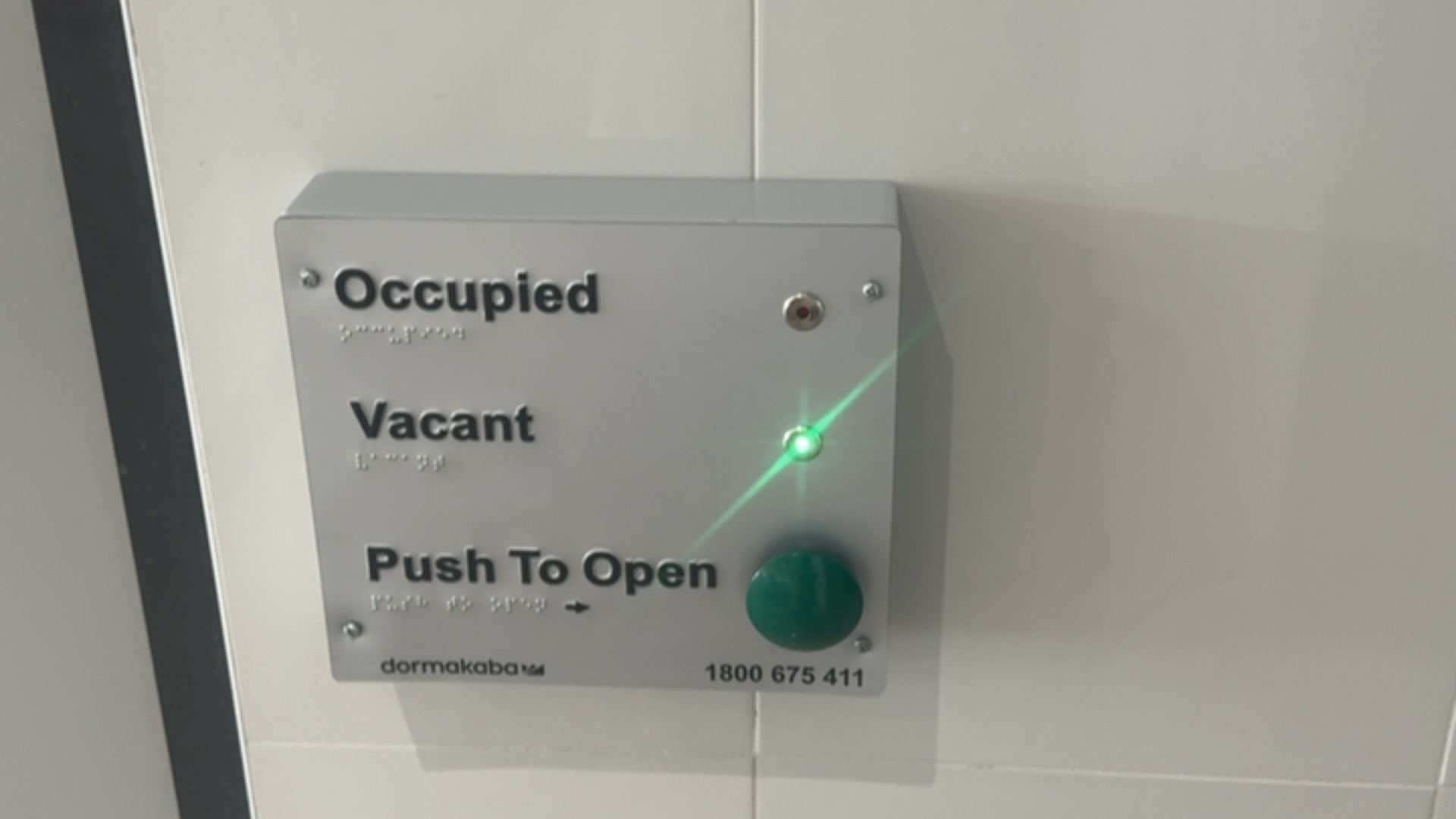 Silver wall-mounted panel with 'Occupied', 'Vacant', and 'Push To Open' text and Braille. A green indicator light is lit next to 'Vacant' above a large green push button.