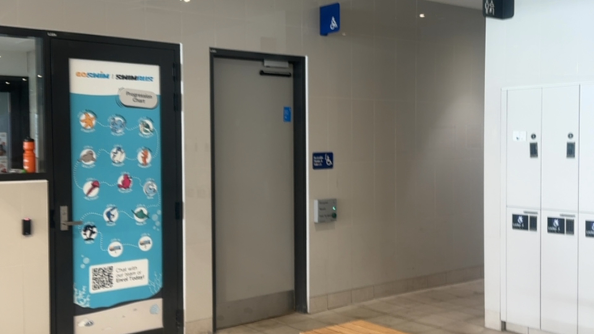 Level tiled floor leads to a grey accessible door with blue wheelchair signs and a push-button automatic opener. A well-lit corridor with white lockers and toilet signs is to the right.