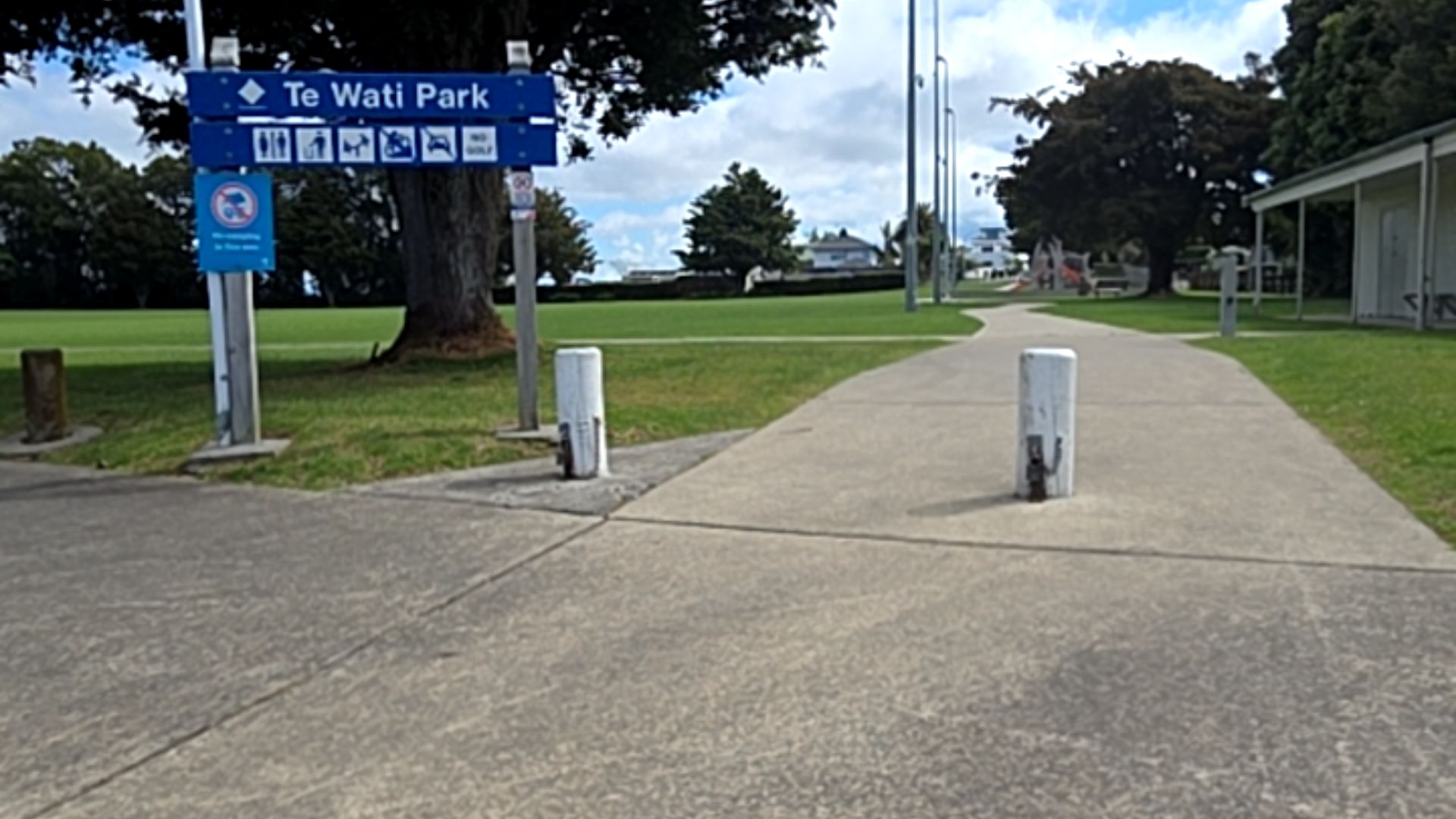Wide, level concrete pathway entrance to Te Wati Park between two white bollards. Blue signage on the left with park icons. Flat grassed areas and a distant playground surround the path.