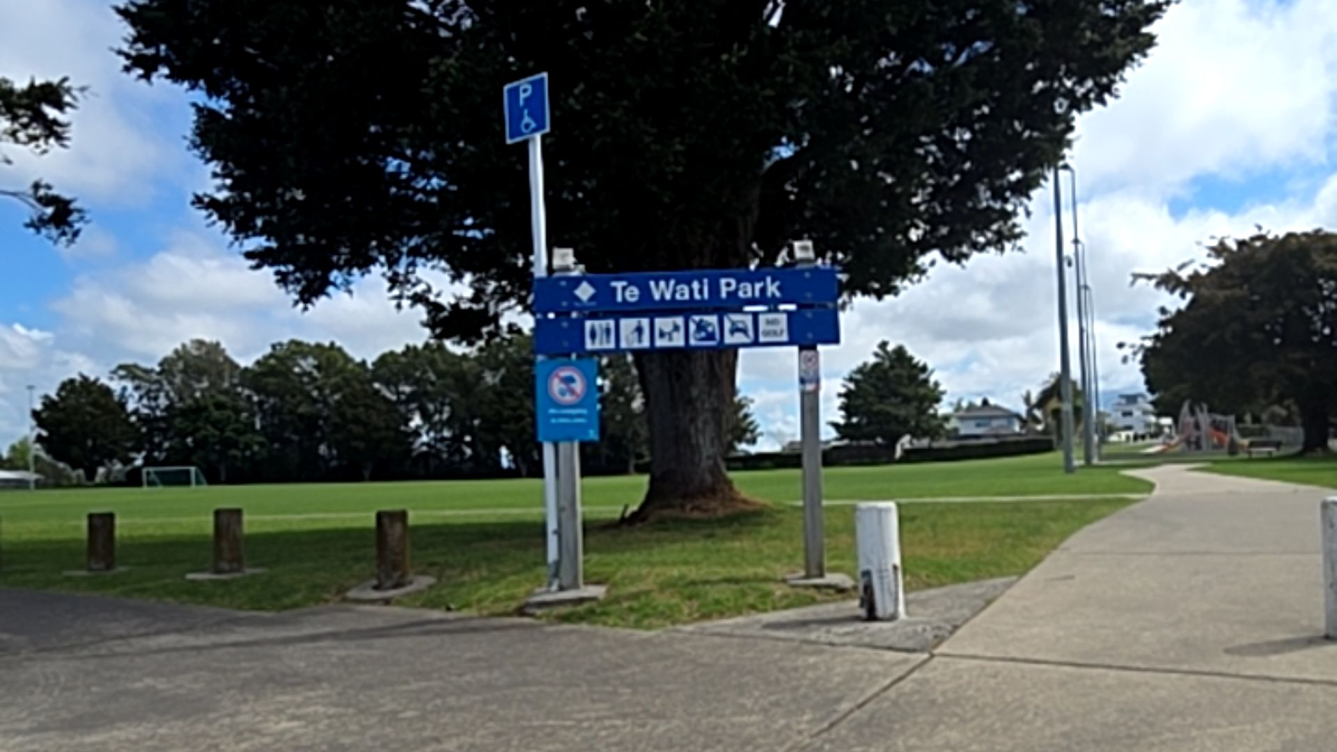 Te Wati Park sign with facility icons and blue accessible parking symbol. Wide, level concrete pathway leads past a large tree toward an open green grassy field with wooden bollards.