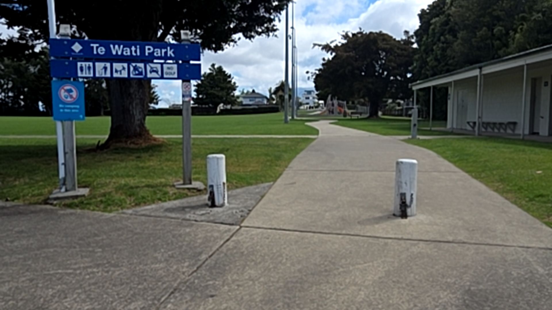 Wide level concrete path enters Te Wati Park between white bollards. A blue signpost with icons is next to a large tree. On the right, a building has a covered porch and outdoor bench seating.
