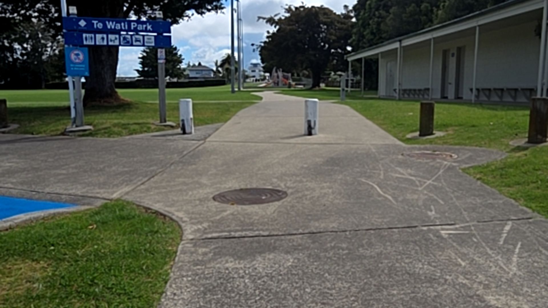 Level concrete path enters Te Wati Park past two white bollards. A blue sign includes accessibility symbols. A building with a flat entrance and covered walkway is on the right.