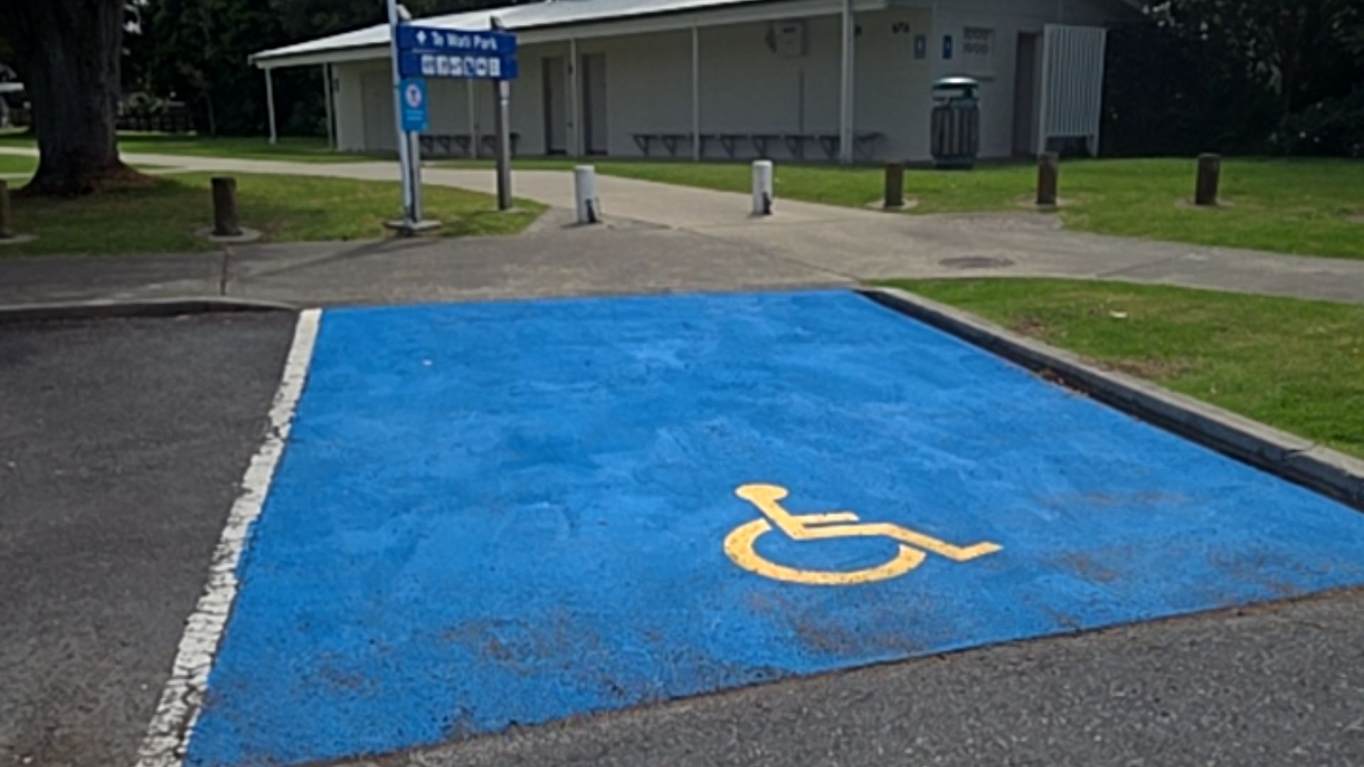 Blue-coloured accessible parking bay with yellow symbol on level asphalt. A flat concrete path leads to a white building with blue signage and bench seating under a covered porch.