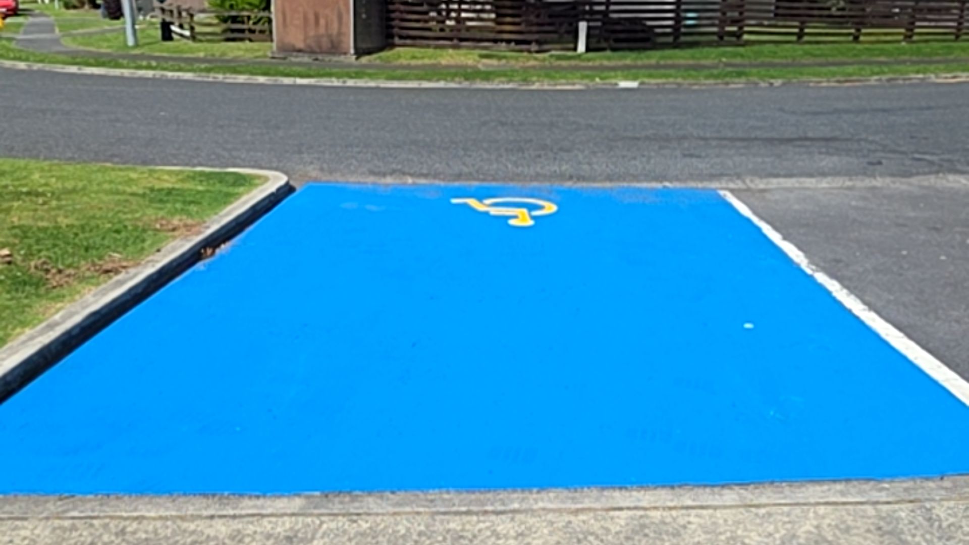 Level accessible parking space painted bright blue with a yellow wheelchair symbol in the centre, bordered by a concrete kerb and a white line on an asphalt surface.