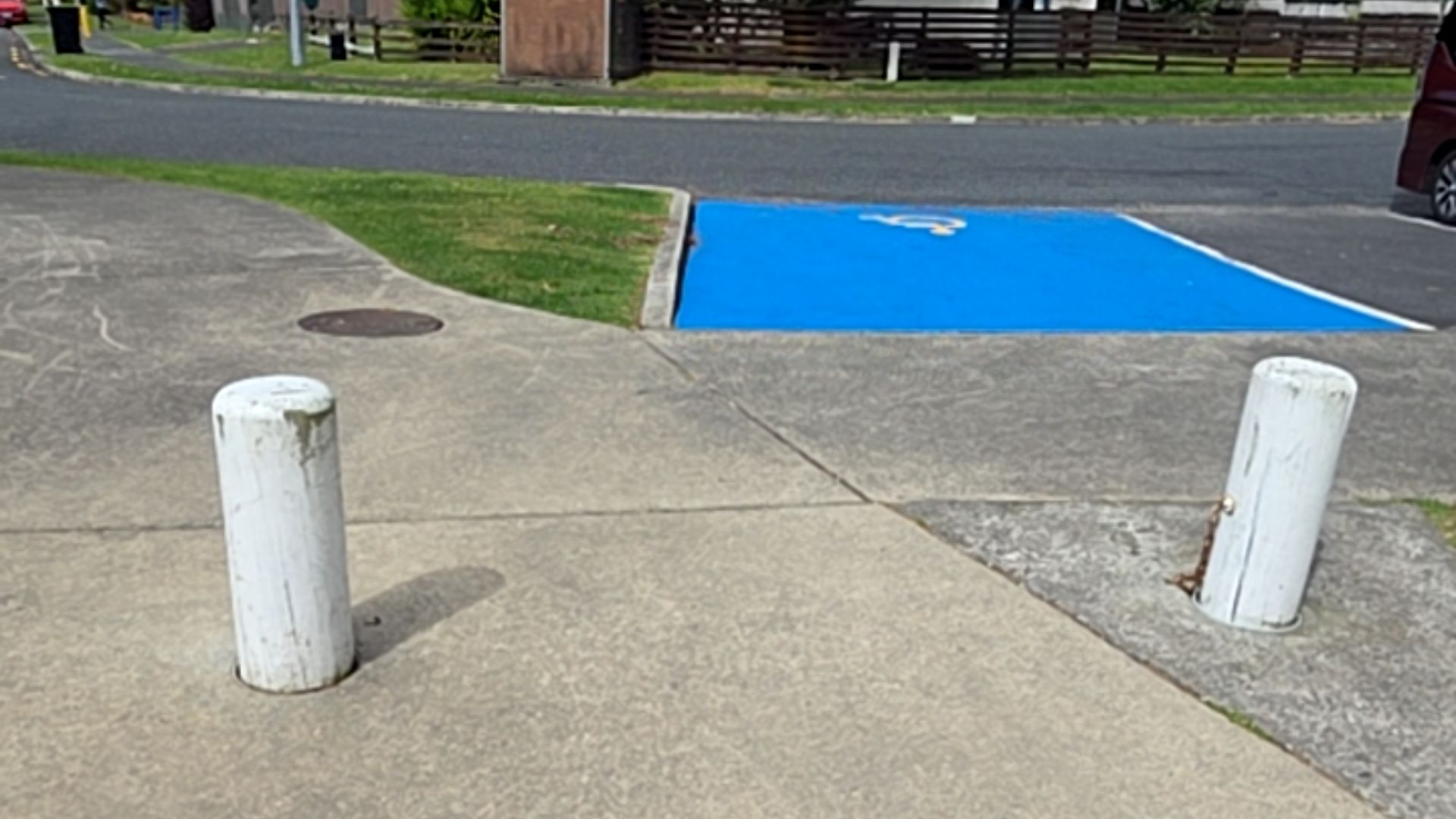 Level concrete path between two white bollards leads to a blue-painted accessible parking space with a white symbol. A concrete kerb ramp connects the path to the asphalt road.