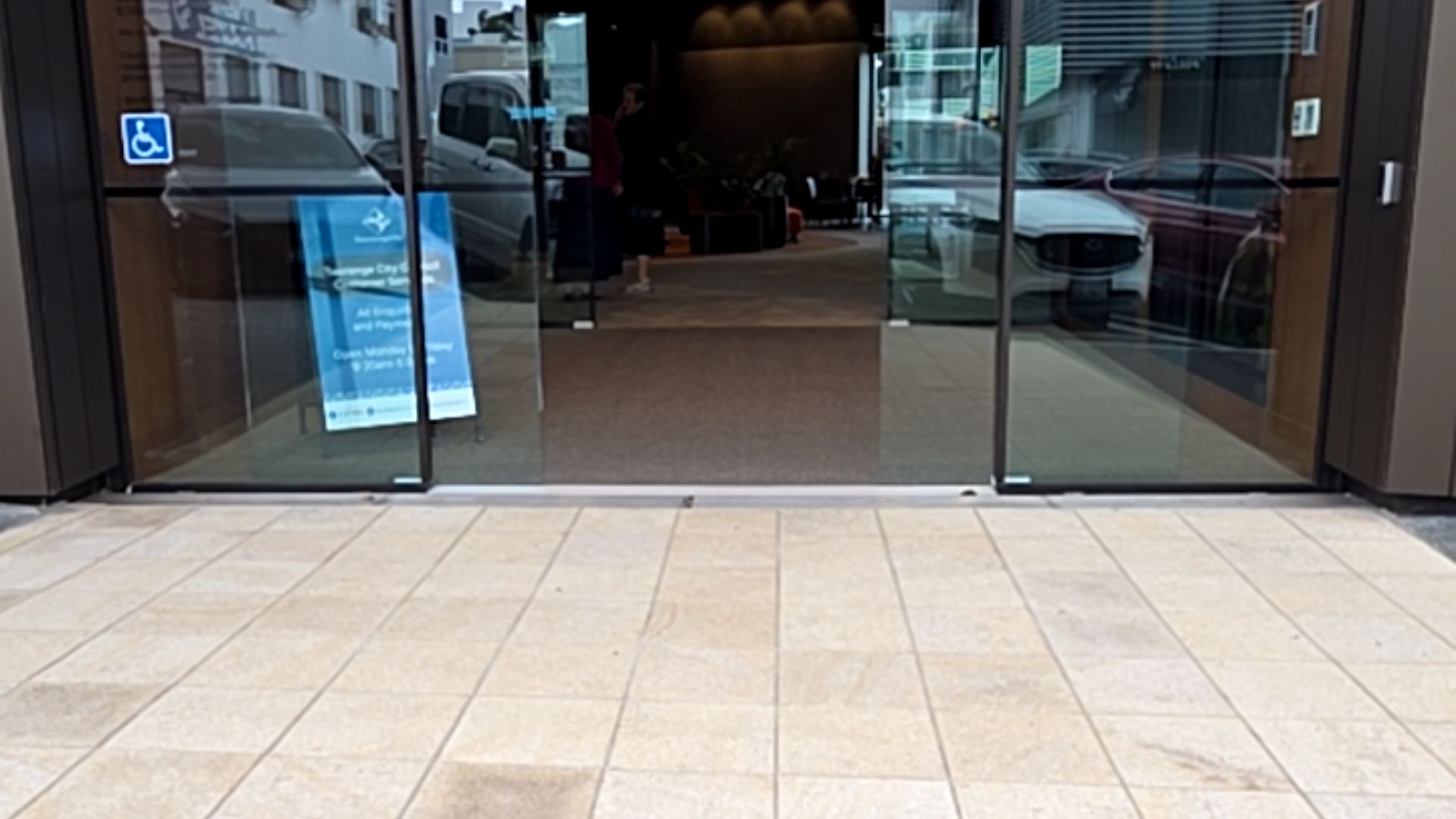 Level entrance with wide automatic sliding glass doors. Light-coloured stone tiles lead to a flush threshold and dark indoor mat. A blue accessibility symbol is on the left glass door panel.