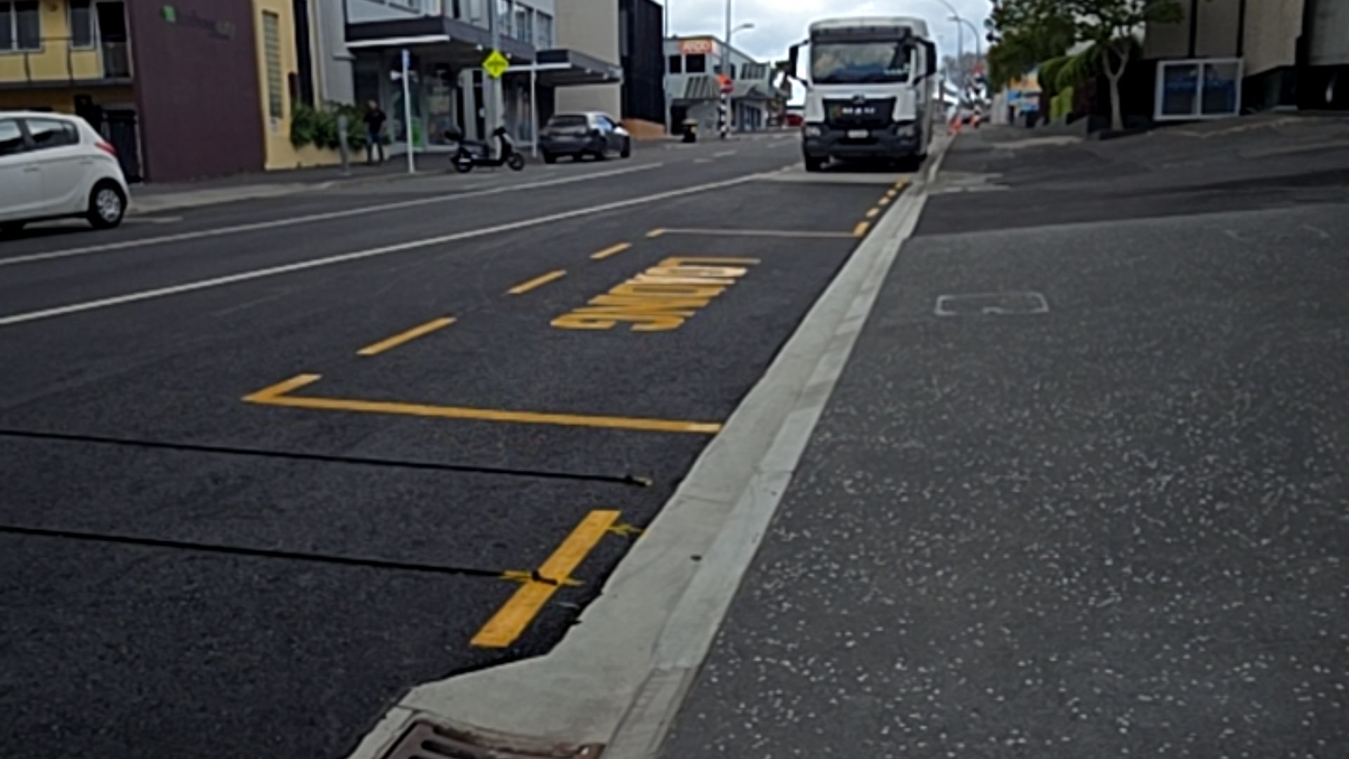 Asphalt road with yellow 'LOADING' signage beside a concrete kerb and level bitumen footpath. Urban buildings and a white truck are visible in the distance.