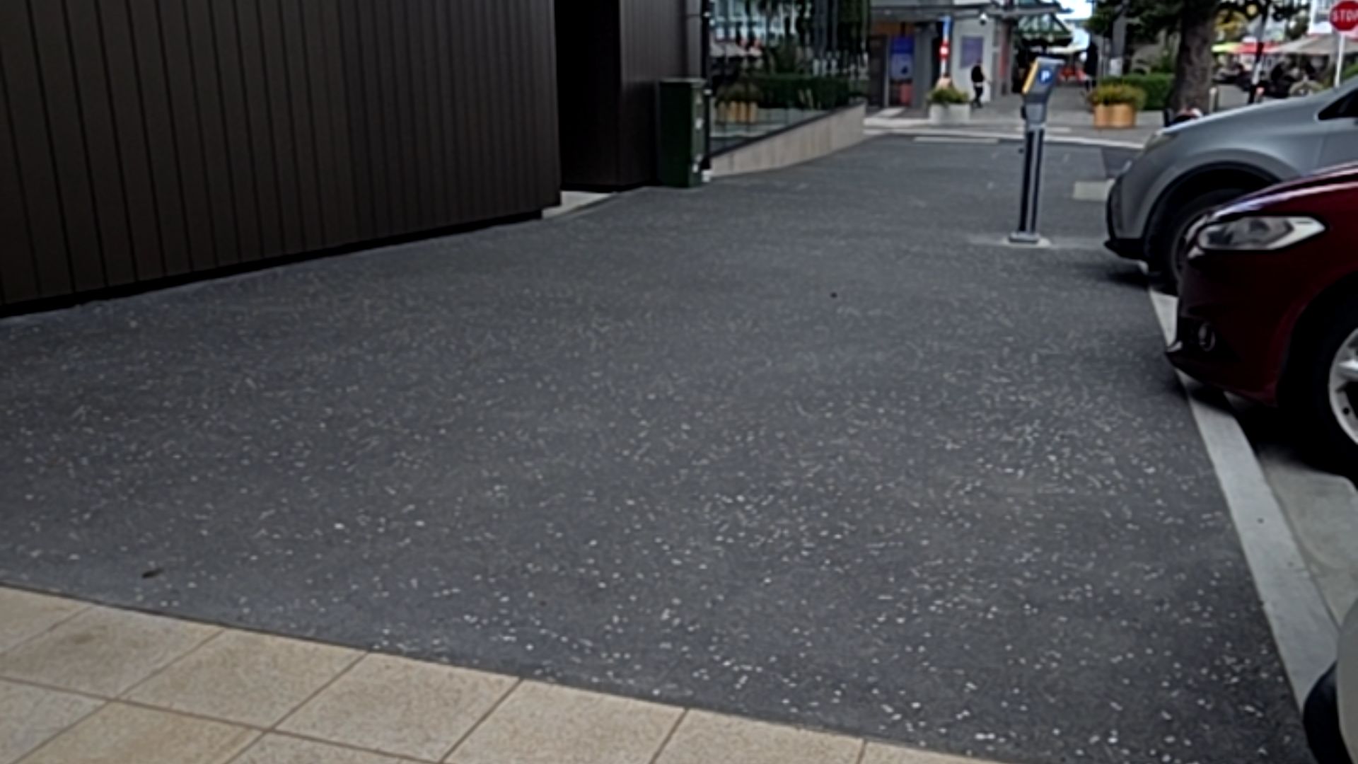 Wide, level dark grey speckled pathway with a smooth transition from light-coloured tiles. A dark brown ribbed wall is on the left, and cars are parked on the right alongside a white line.