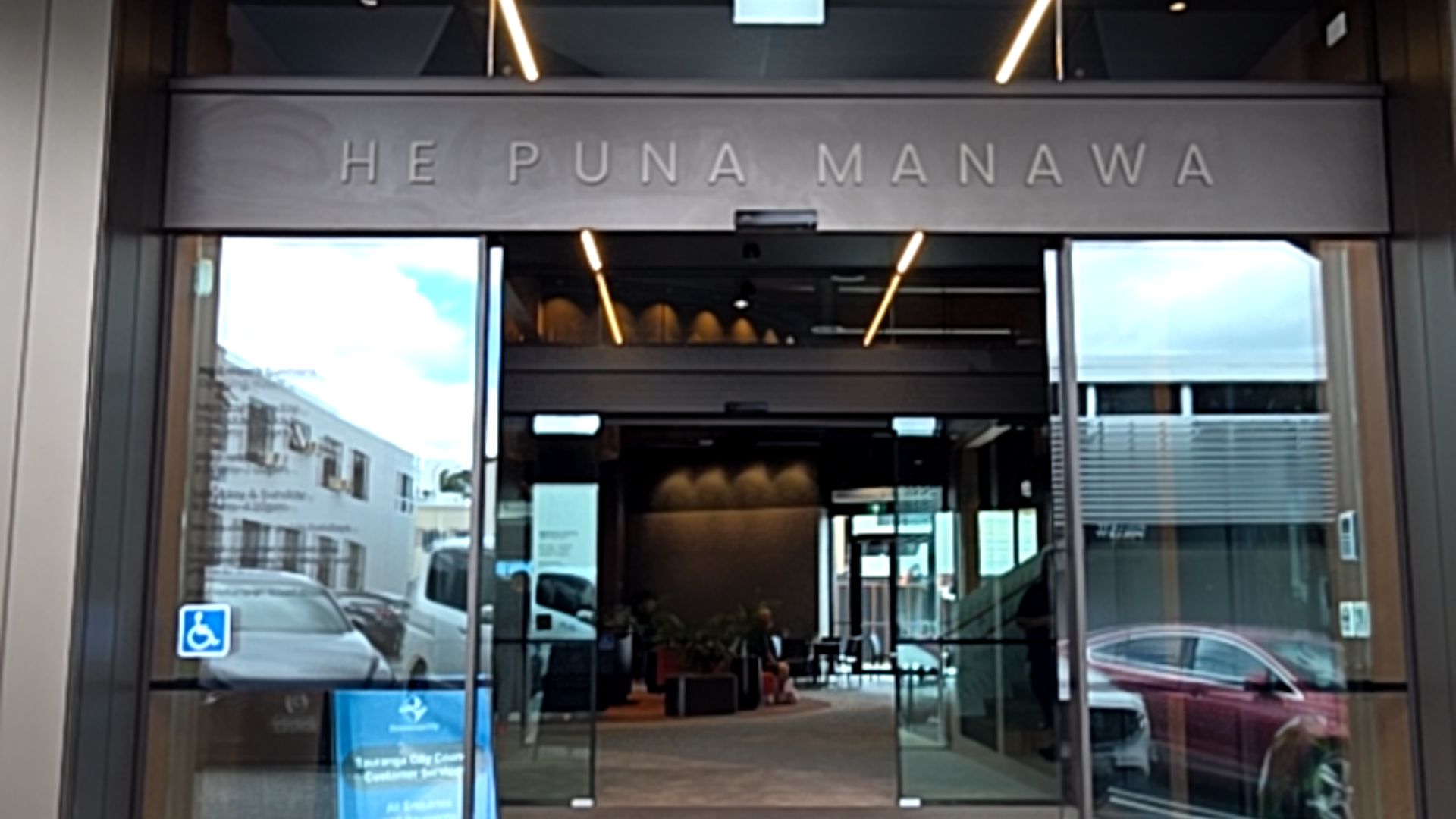 Level entrance with automatic sliding glass doors. Large 'HE PUNA MANAWA' sign above and blue accessibility symbol on glass. Dark-coloured flat floor leads inside under warm strip lighting.