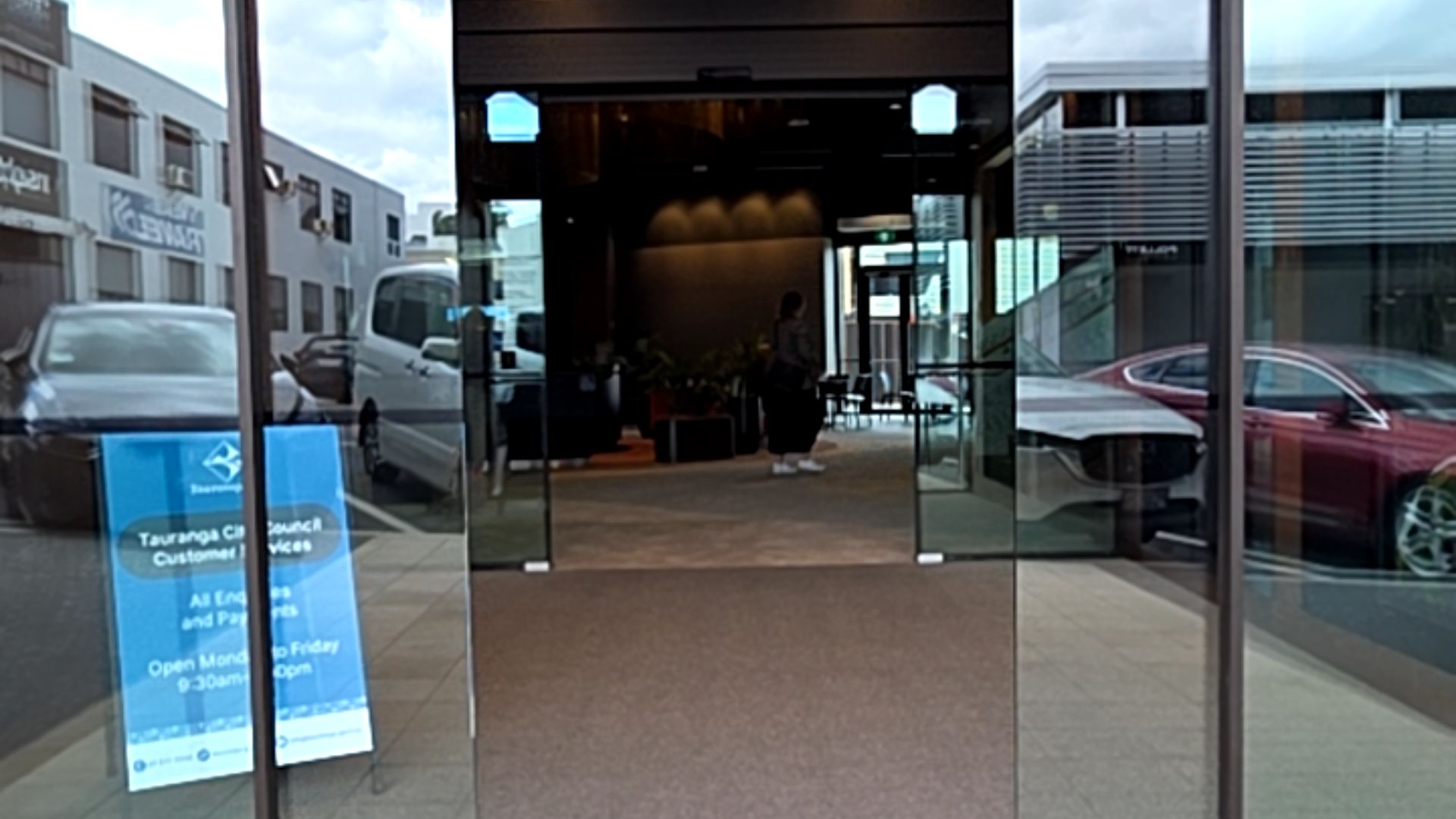 Automatic glass sliding doors provide wide, level access from a light-grey paved pathway to a dark carpeted interior. A blue sign with white text is positioned on the left of the entrance.