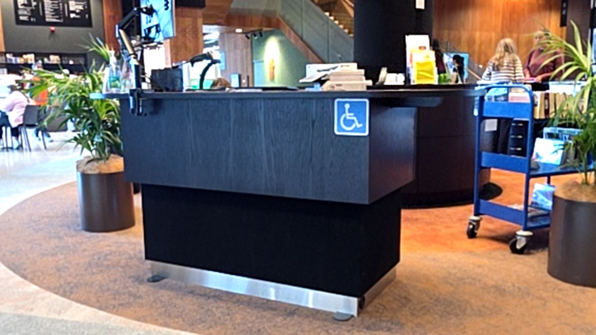 Black timber service desk with a lowered counter section marked by a blue and white wheelchair symbol. The desk sits on level, tan and grey textured carpet.