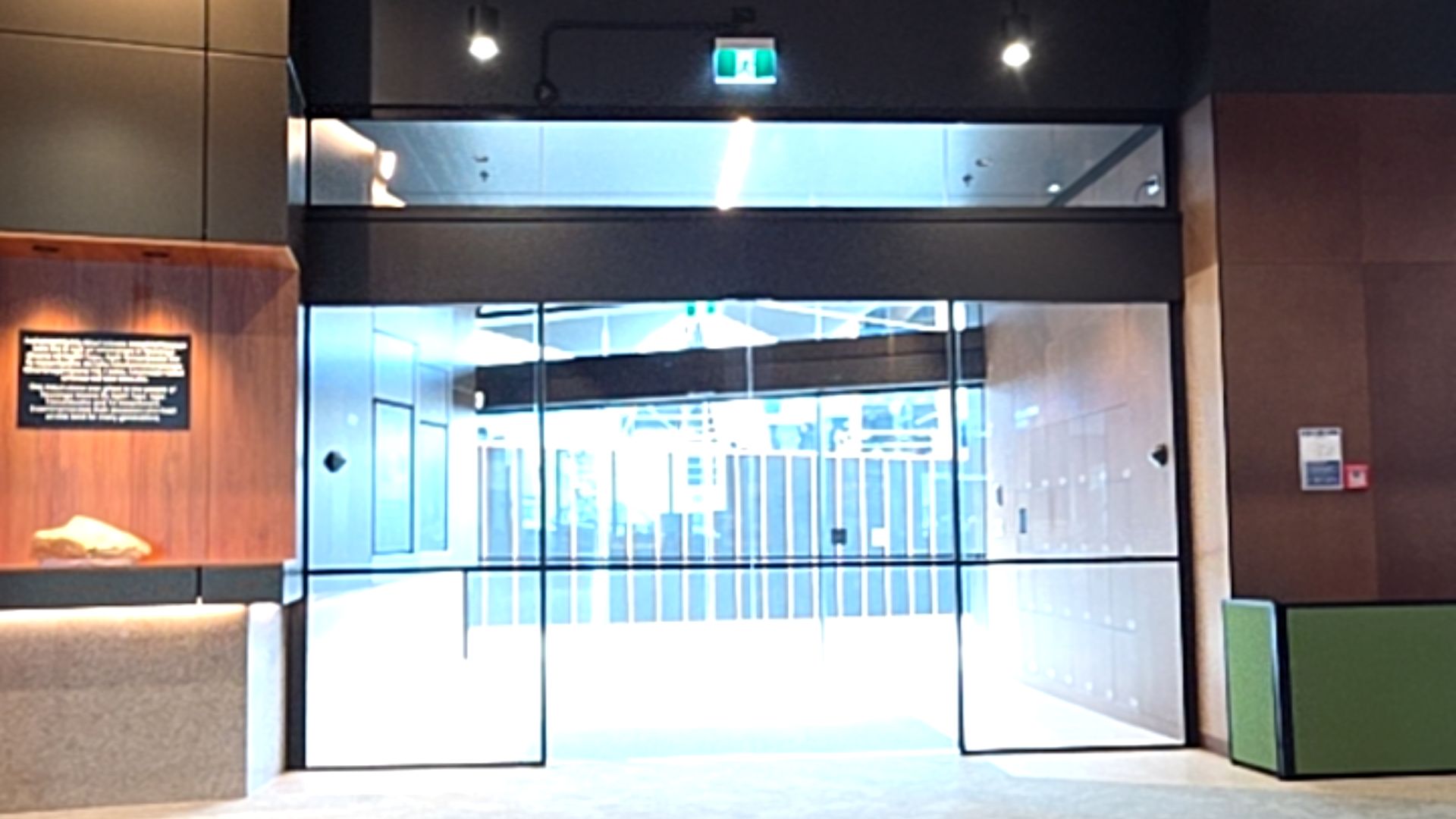 Wide level entrance with automatic double glass sliding doors and flat, light-coloured flooring. Brightly lit interior beyond. A green illuminated exit sign is above the dark door frame.