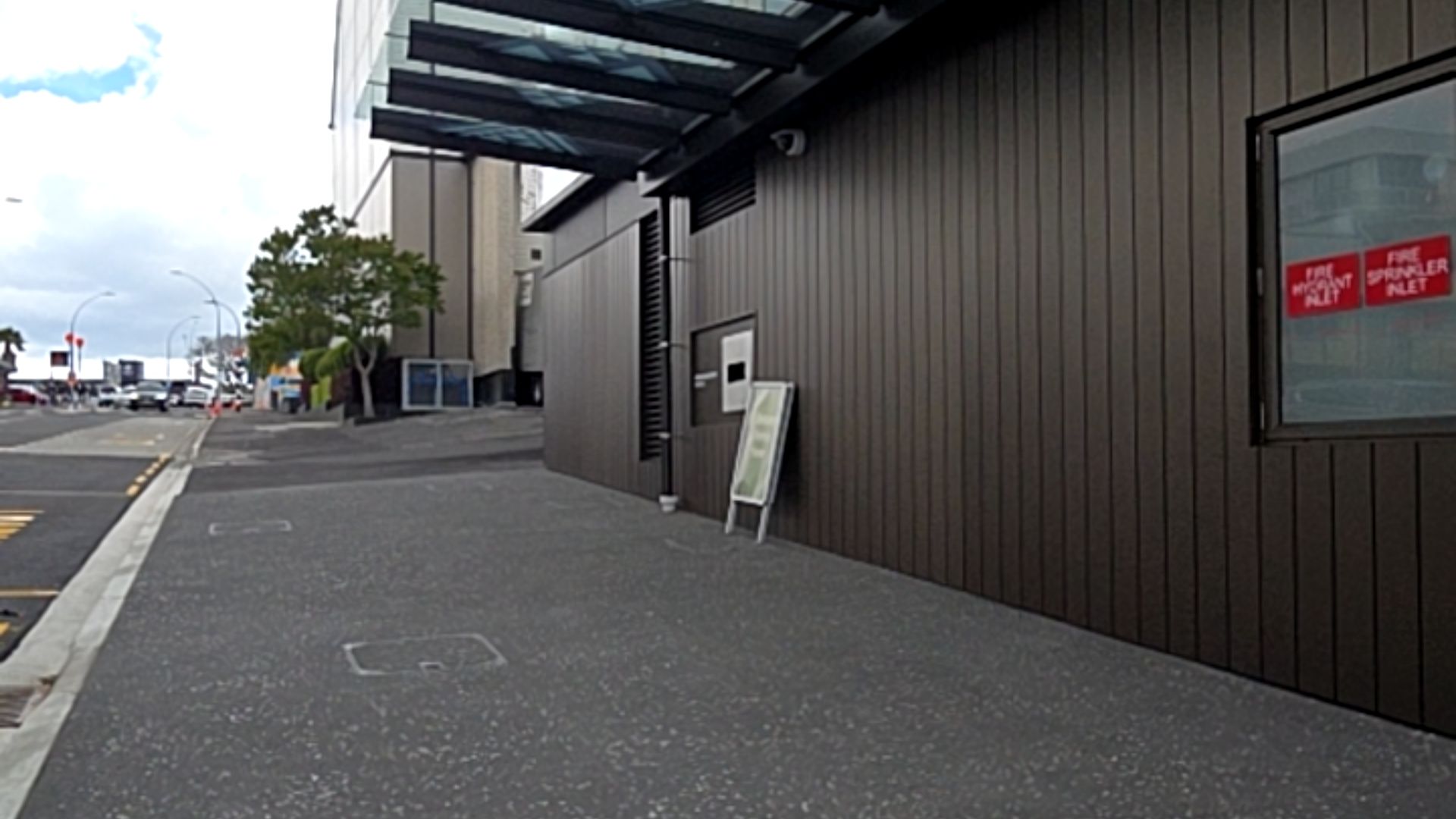 Wide, level grey asphalt pathway beside a dark brown vertically-panelled wall with a glass canopy overhead. A white A-frame sign stands on the path near a recessed section.