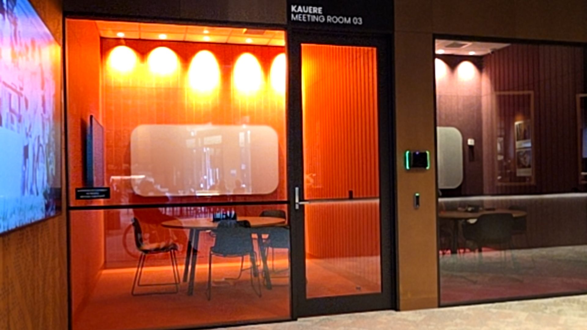 Level entrance to orange-coloured meeting rooms with glass walls. Wide door with lever handle and card reader. Interior features a round table and chairs on a flat, even floor surface.
