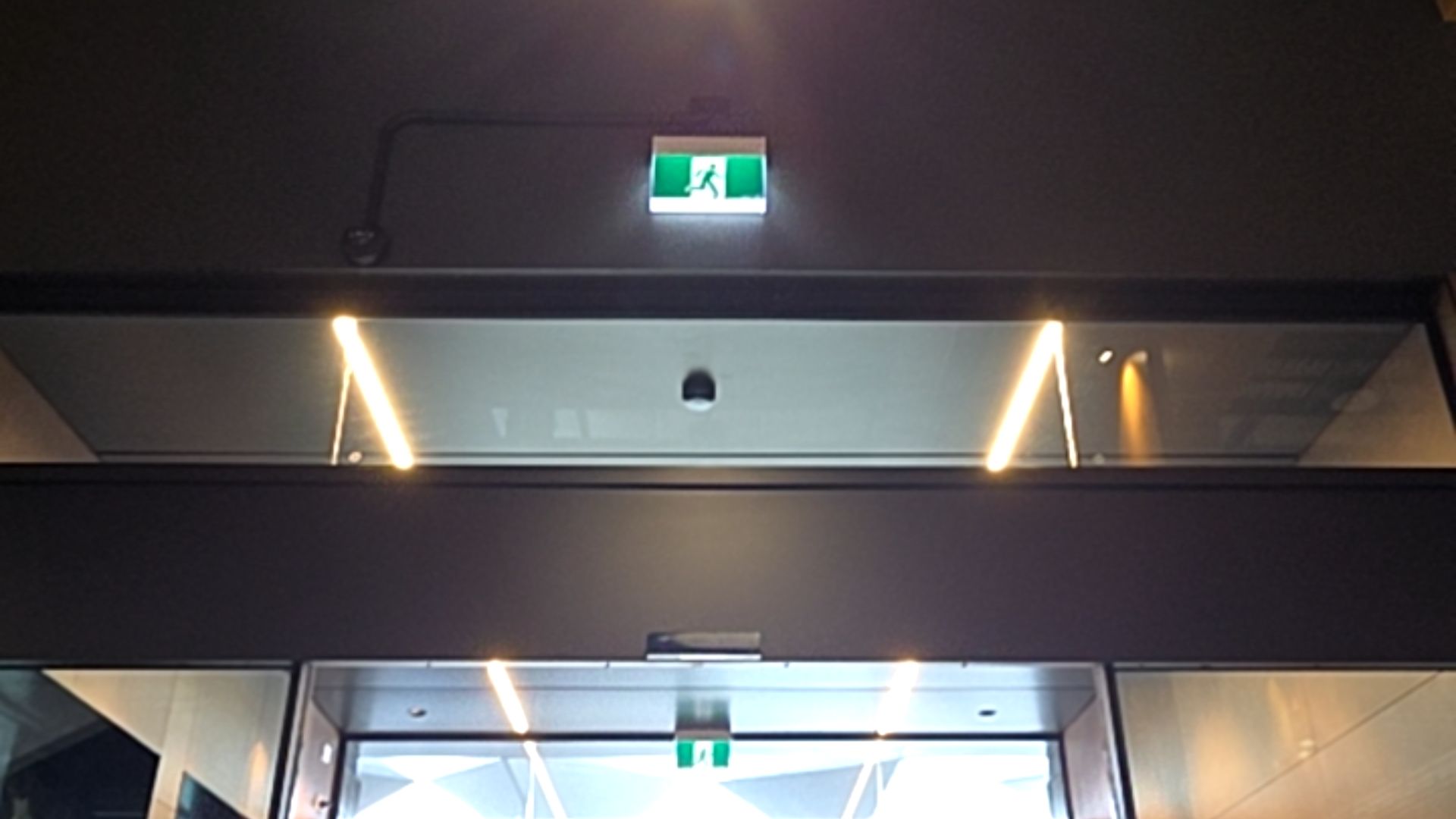 Automatic glass sliding doors with a level entrance. A green illuminated exit sign is mounted above the dark grey door frame. Two vertical strip lights illuminate the glass section above the doors.