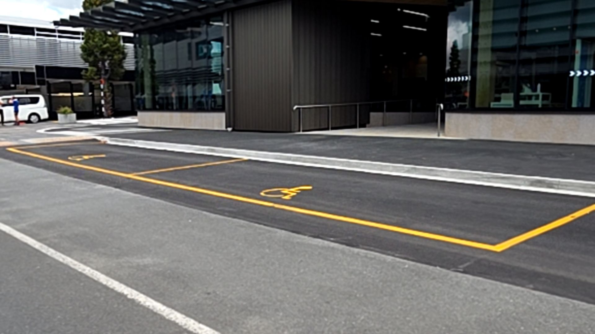 Two accessible parking spaces with yellow lines and symbols on dark asphalt. Level concrete pathway leads to a building entrance with silver handrail and dark grey walls.