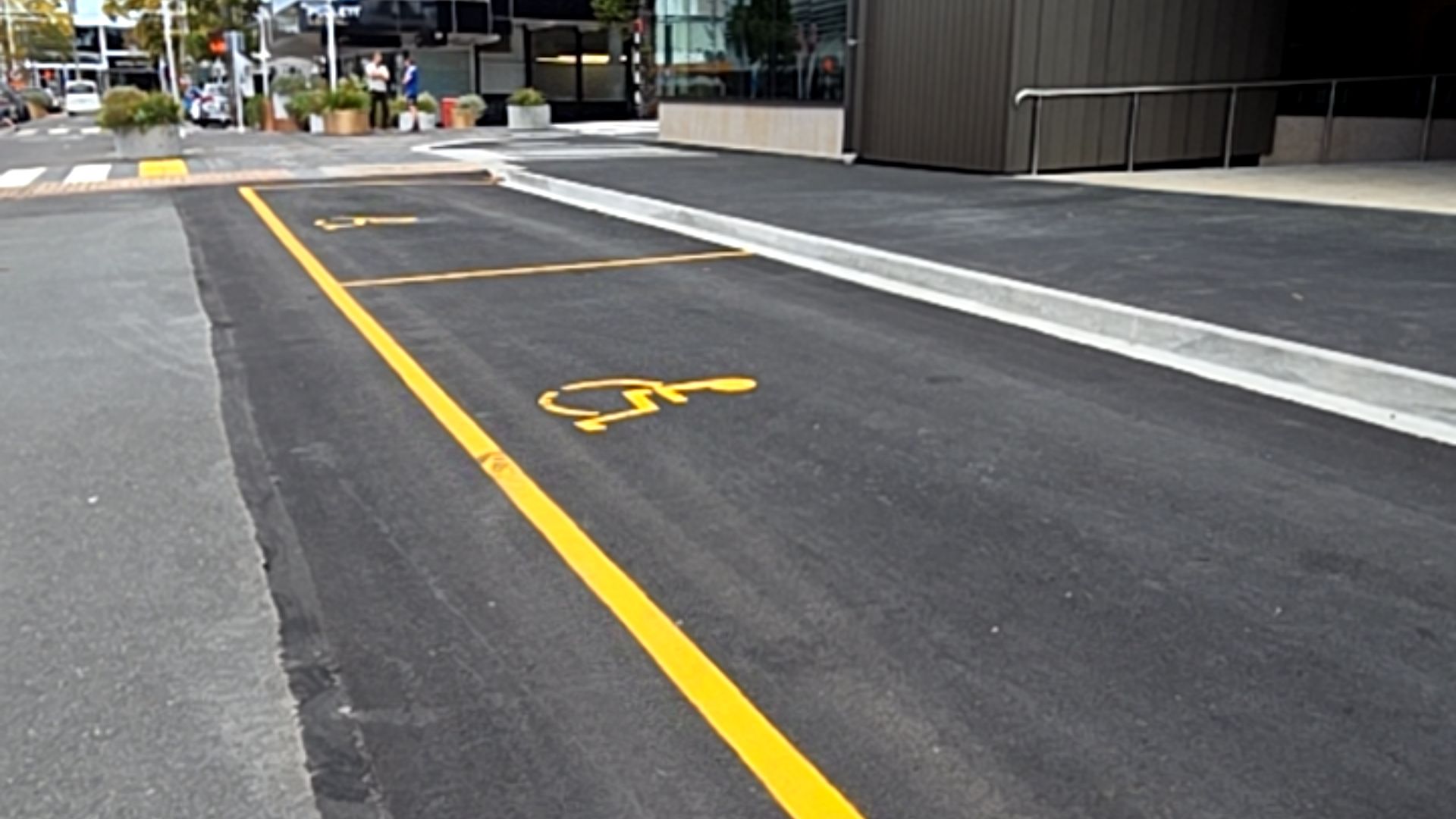 Two accessible parking spaces on level black asphalt, marked with yellow wheelchair symbols and lines. A concrete kerb borders the spaces, adjacent to a wide, level paved area.