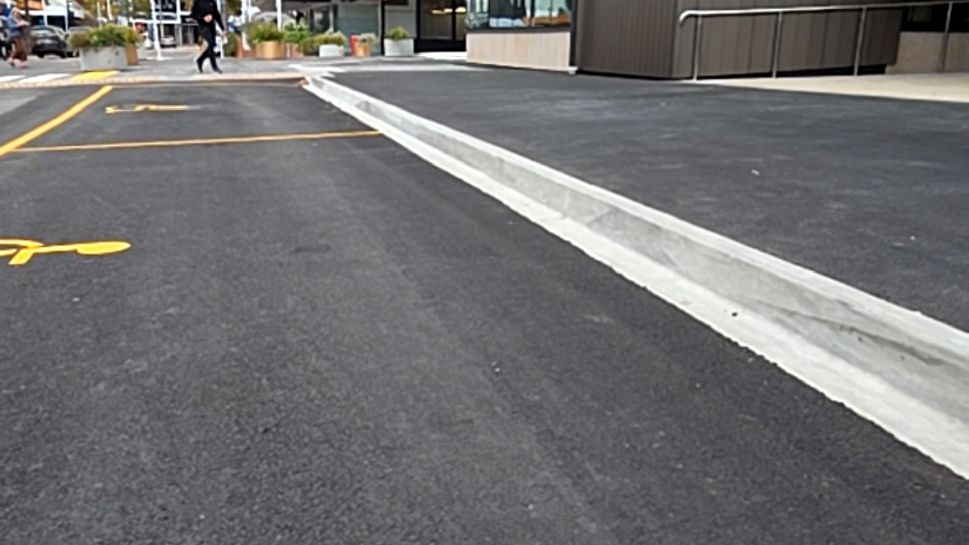 Accessible parking spaces marked with yellow symbols on dark, level asphalt. A concrete gutter borders the area. Metal handrails line a building entrance in the background.
