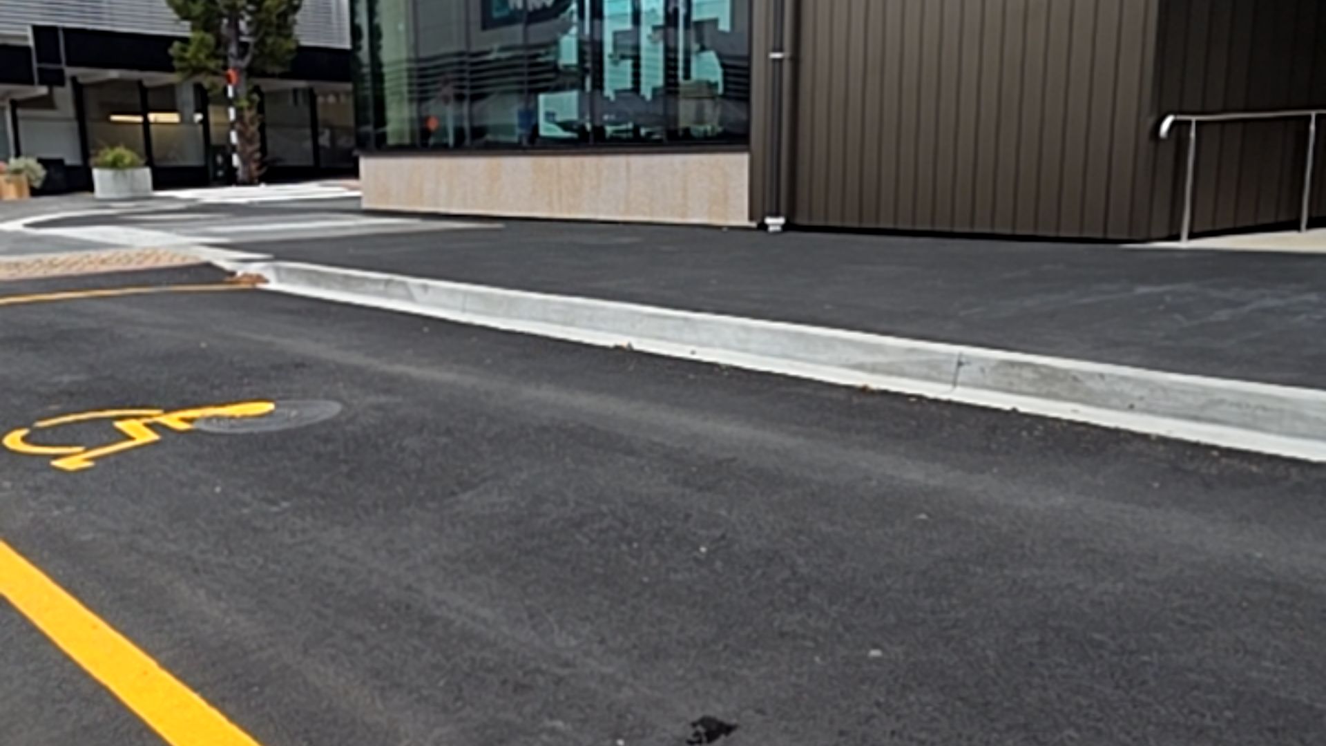 Accessible parking space with yellow symbol on asphalt. A concrete kerb borders a level pathway leading to a building entrance with a metal handrail.