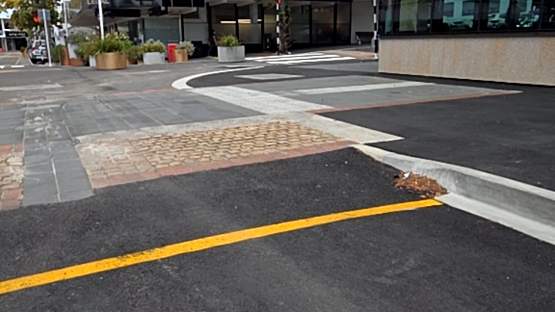 Concrete kerb ramp connects black asphalt with a yellow line to a multi-textured pathway of grey stone pavers and brown bricks. Level access point in natural daylight.