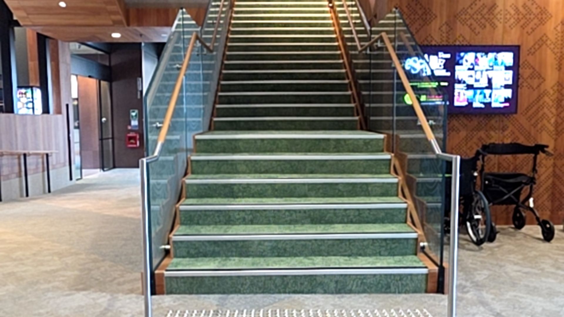 Green carpeted stairs with metal nosing and tactile studs at the base. Glass balustrades have wooden handrails. A mobility walker is parked on the level carpeted floor to the right.