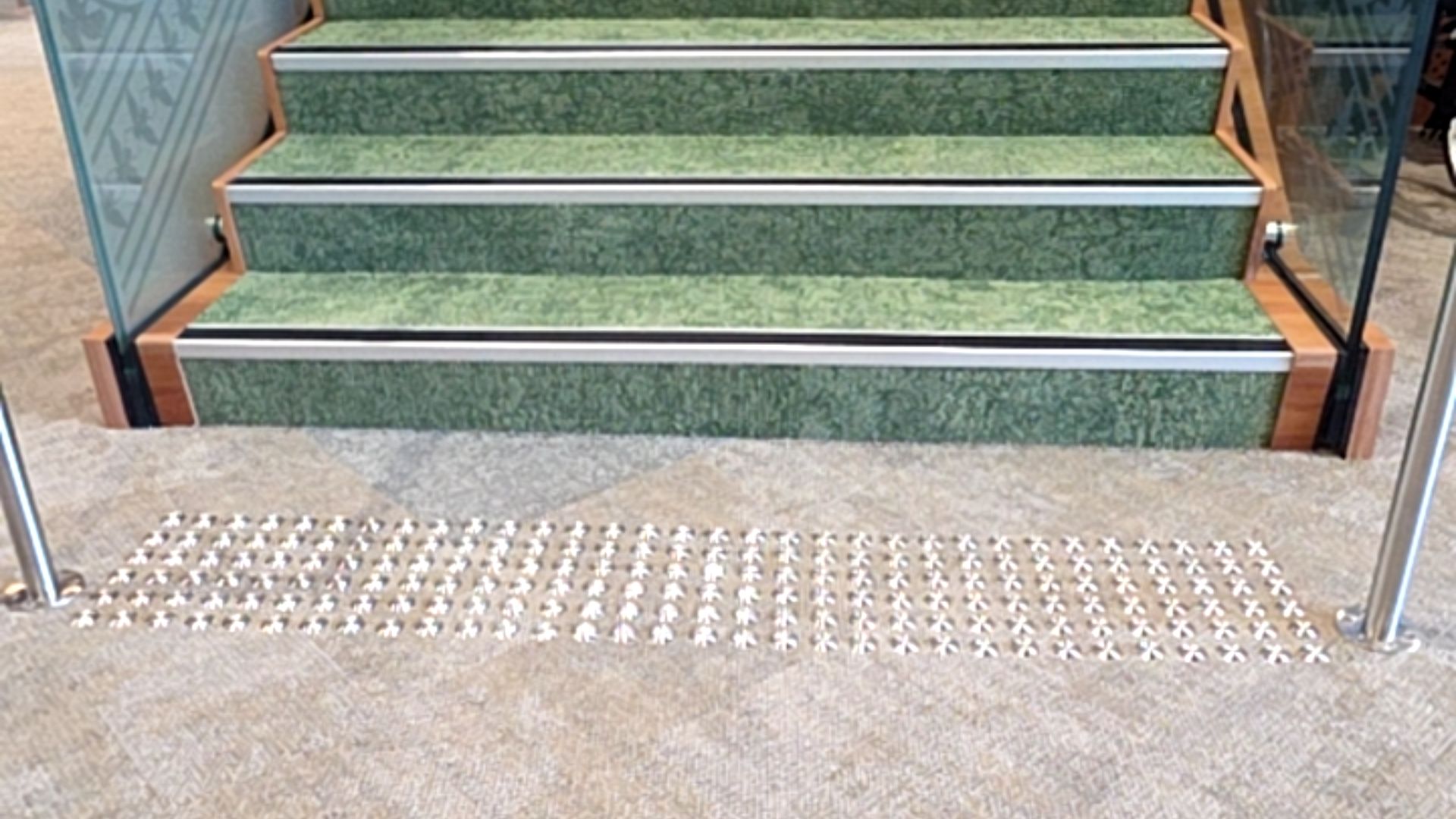 Green carpeted stairs with silver and black nosing. A grid of silver tactile studs is on the grey carpet at the base. Silver metal handrails are on both sides.