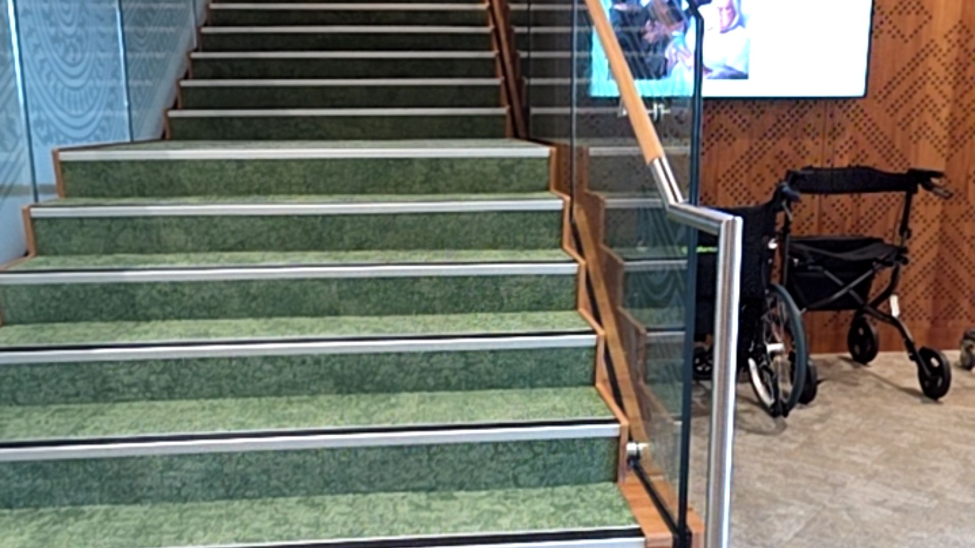 Green carpeted stairs with high-contrast silver nosing and handrails. A wheelchair and walker are parked on the level floor at the base of the stairs beside a patterned timber wall.