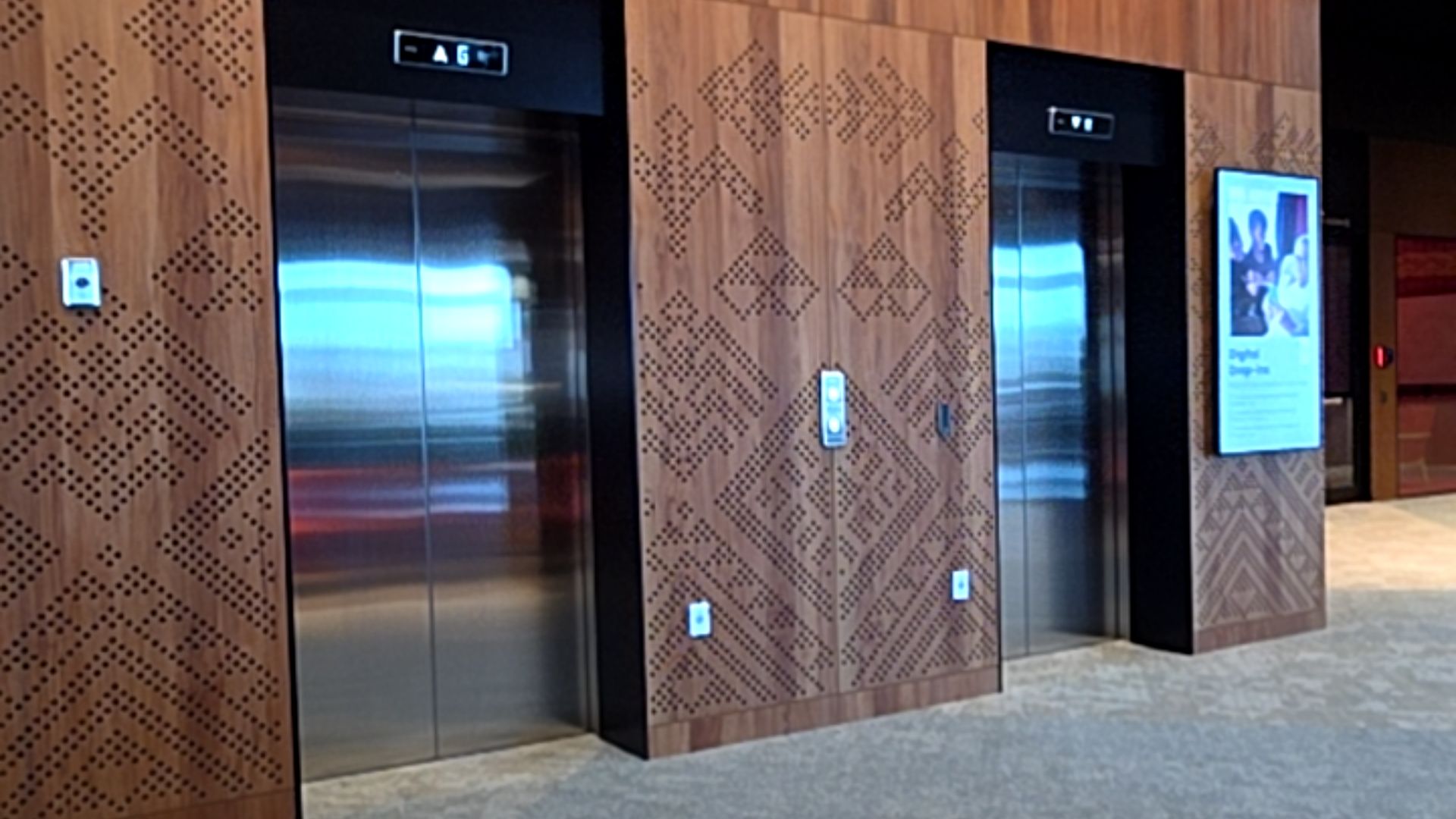 Two stainless steel lifts set into timber-panelled walls with perforated patterns. Level grey carpeted floor. Wall-mounted call buttons and digital floor indicators above the doors.