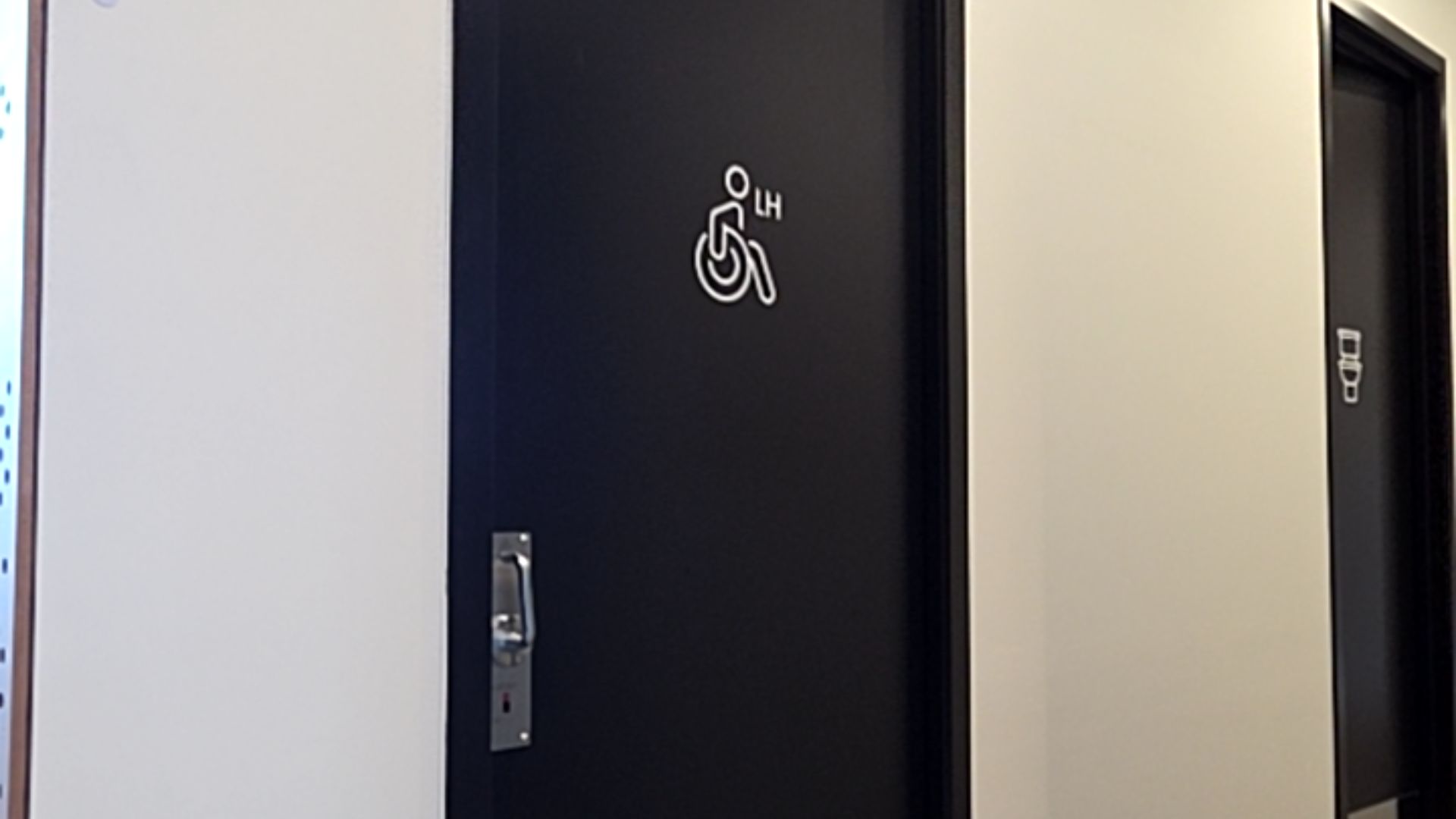 Black door with white wheelchair symbol and 'LH' text. Another black door to the right shows a white toilet icon. Doors are set in white walls with level floor entry.