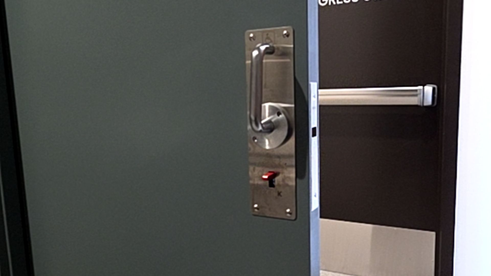Grey door with stainless steel plate featuring a vertical D-handle, embossed wheelchair symbol, and red lock toggle. A dark door with a silver horizontal push bar is visible in the background.