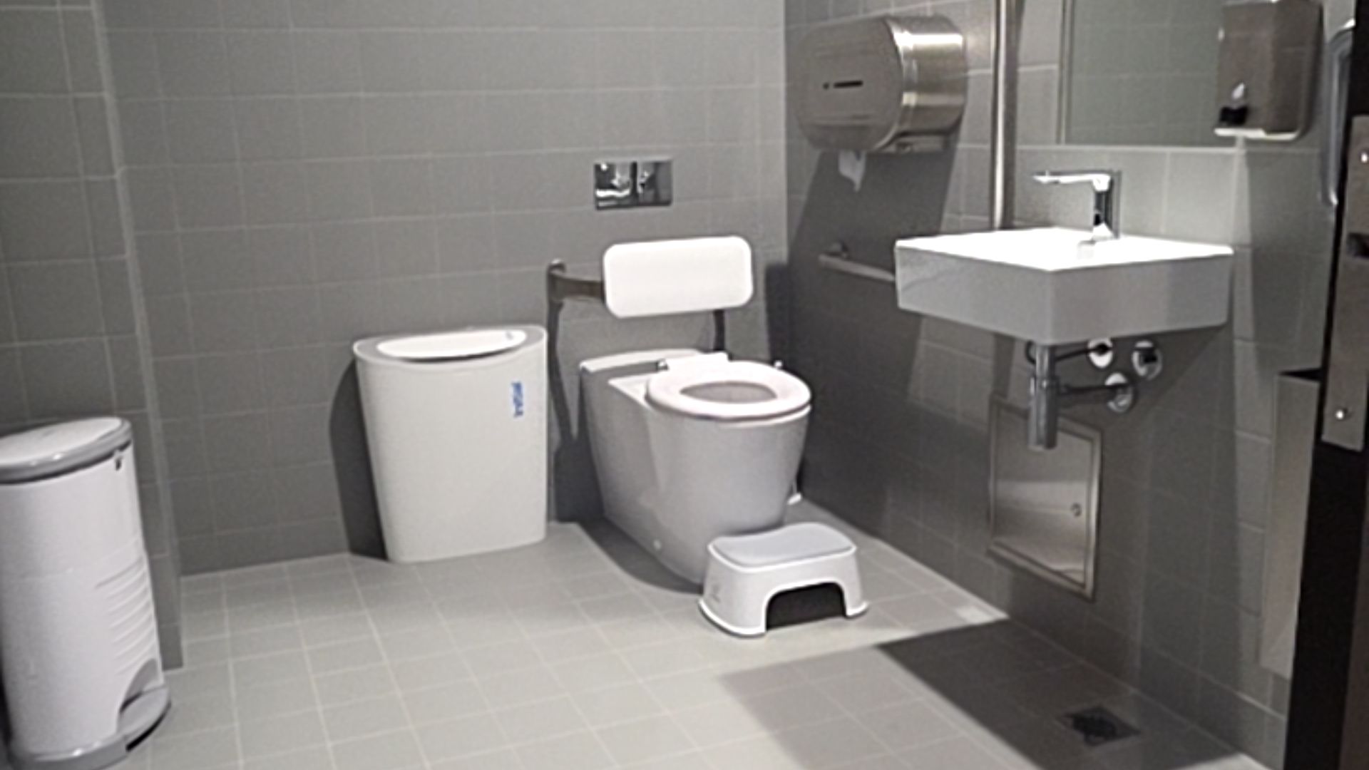 Level grey tiled floor and walls with white toilet and backrest, stainless steel grab bars, and wall-mounted sink with open space underneath and lever tap. Small stool near toilet.
