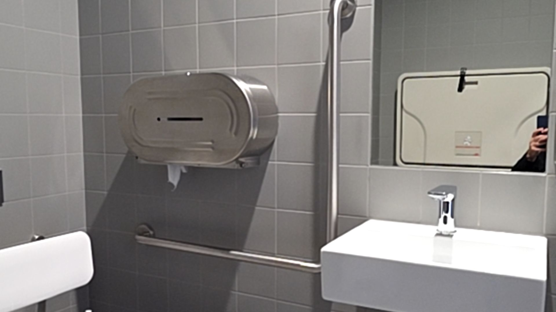 Grey tiled wall with metal grab rails beside a white wall-mounted sink. Large metal dispenser above a horizontal rail. White padded backrest on the left. Mirror above sink reflects changing table.