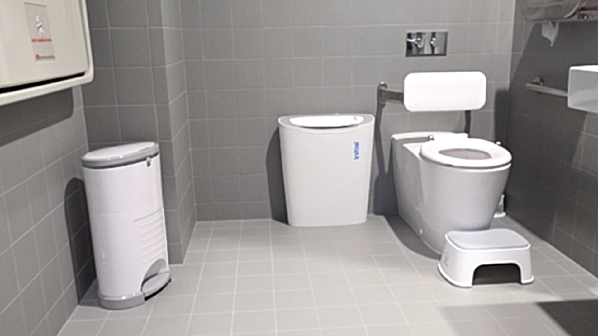 Level grey tiled bathroom. White toilet with padded backrest and silver grab rail. Features a small white step stool, white bins, and a fold-down baby change station on the left.