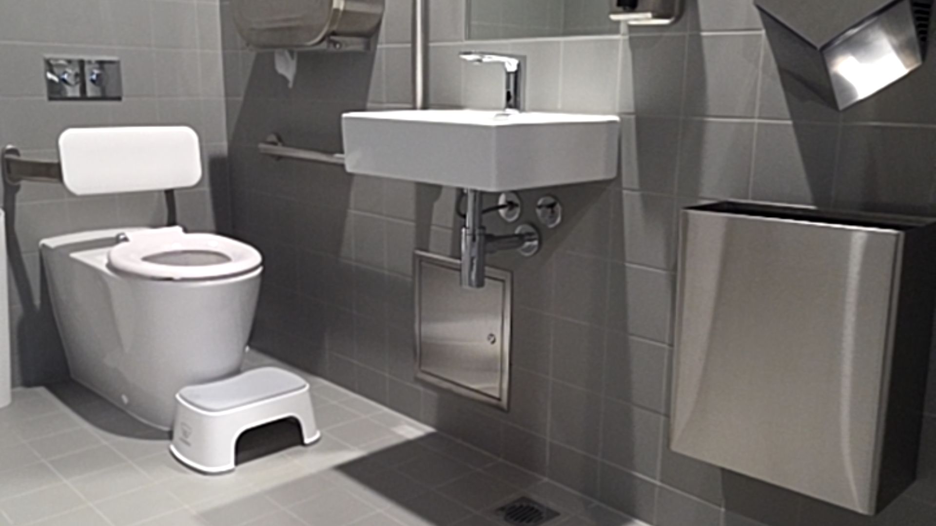 White toilet with backrest and horizontal grab rail. Wall-mounted white basin with lever tap. Grey tiled floor with drain, small step stool, stainless steel waste bin and hand dryer.