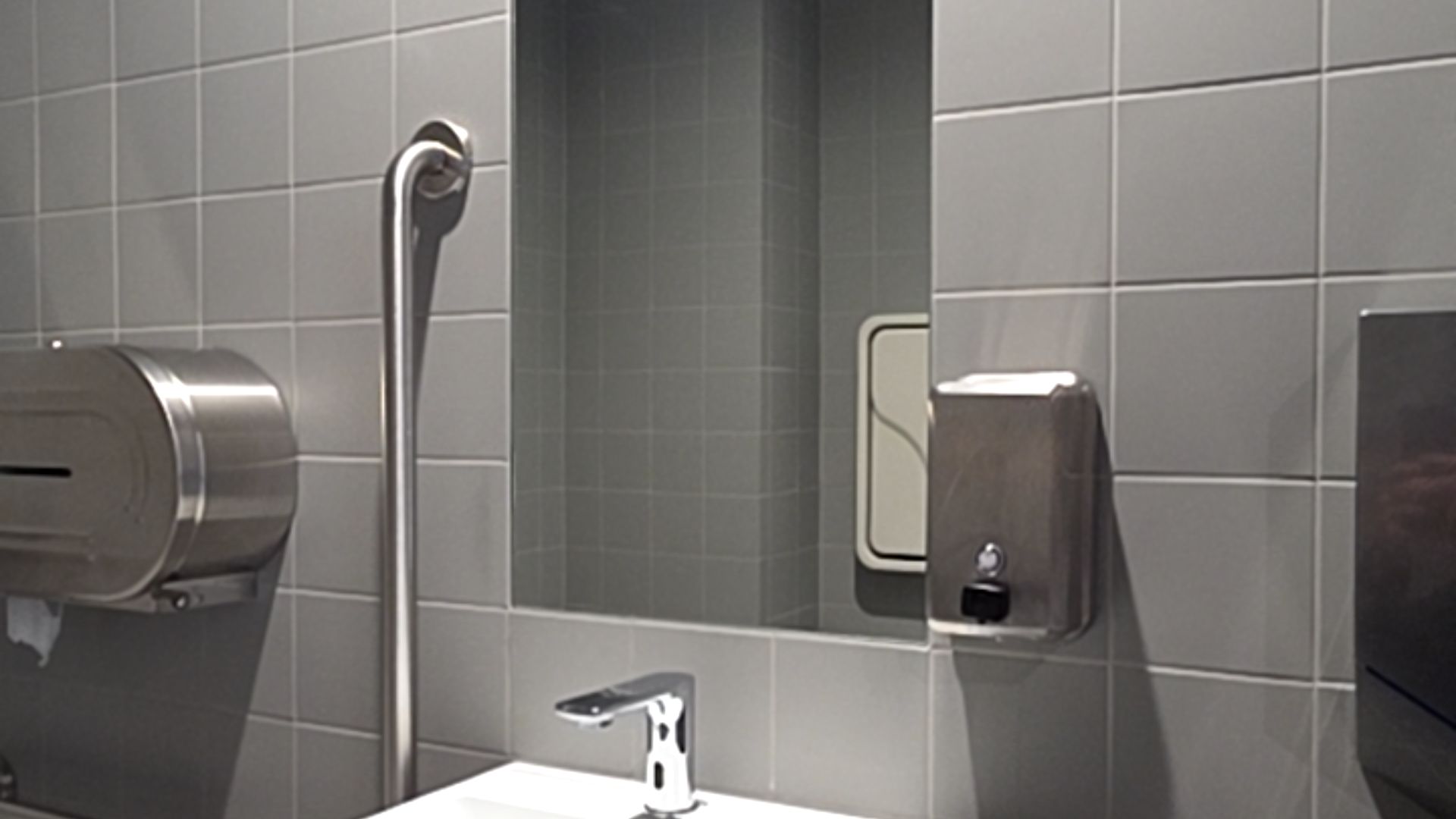 Accessible basin with sensor tap, vertical stainless steel grab bar, and soap dispenser on grey tiled walls. Mirror reflects a wall-mounted baby change table.