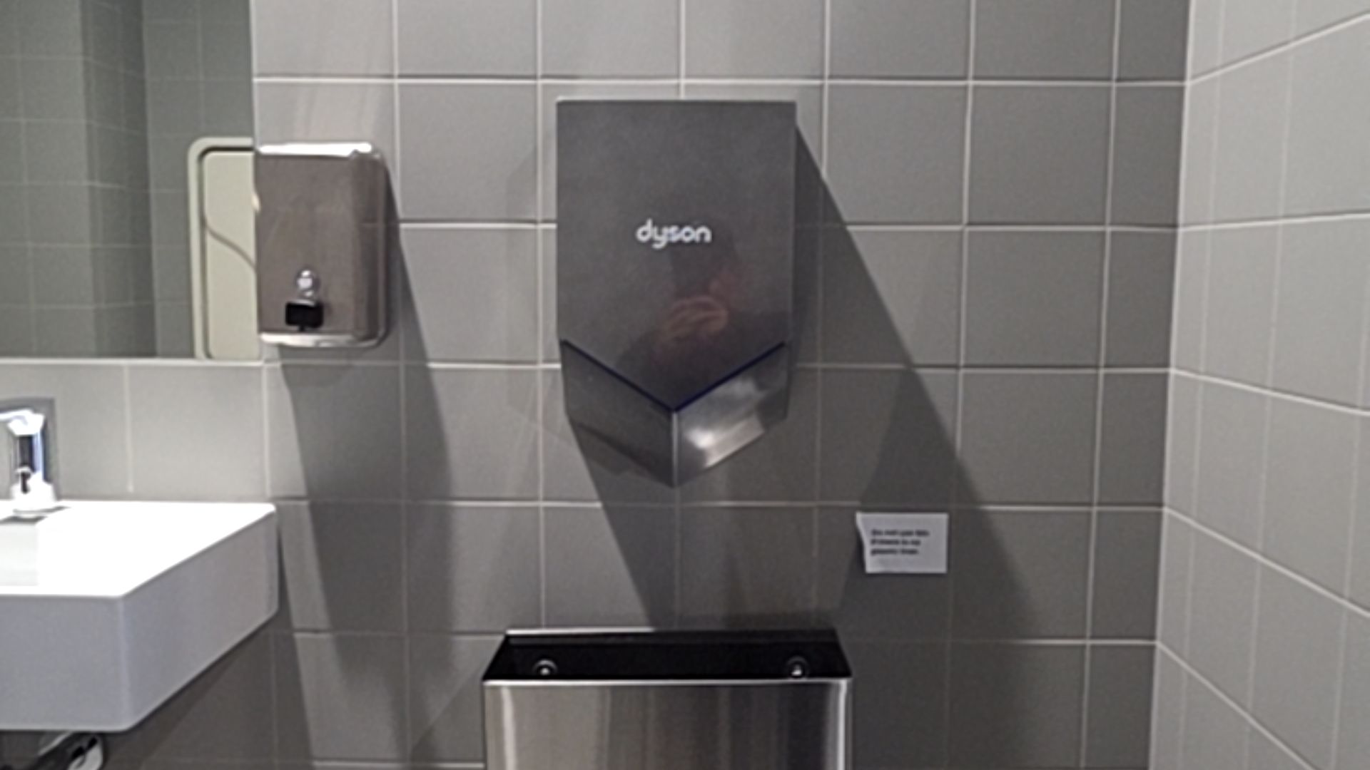 Stainless steel wall-mounted hand dryer and soap dispenser on grey tiled wall. White ceramic sink to left, open-top metal bin below. Small white sign on wall. Level tiled surfaces.