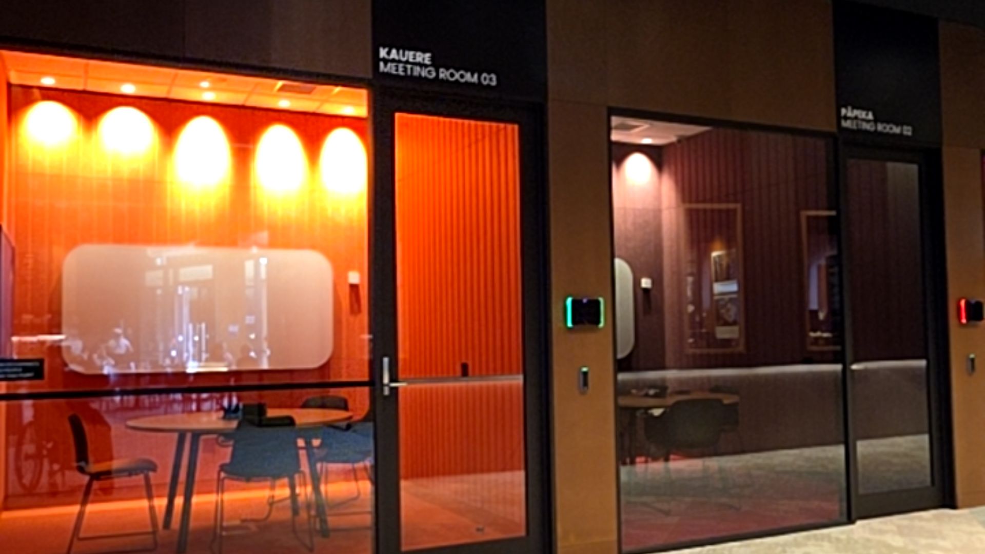 Two glass-walled meeting rooms with level entries. Room 03 has bright orange lighting, a round table and chairs. Overhead signage and digital displays are next to each door.