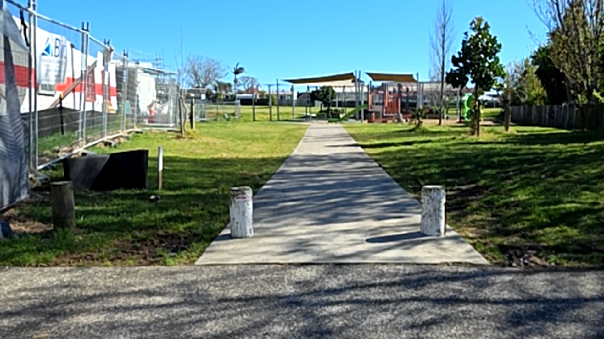 Level concrete path between two white bollards, leading to a playground with shade sails. Grass on both sides and a metal construction fence on the left.