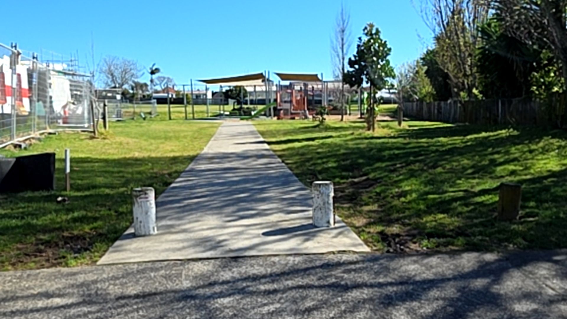 Level concrete pathway starting between two white bollards, leading through a grassy park towards a playground with shade sails. The path is wide, flat, and unobstructed.