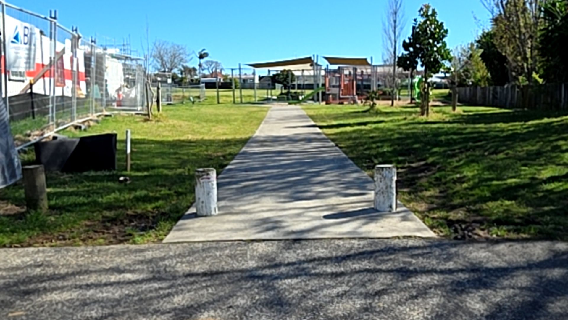 Wide, level concrete pathway starting between two white bollards from an asphalt area. Path leads across flat grass towards a playground with shade sails in the distance.