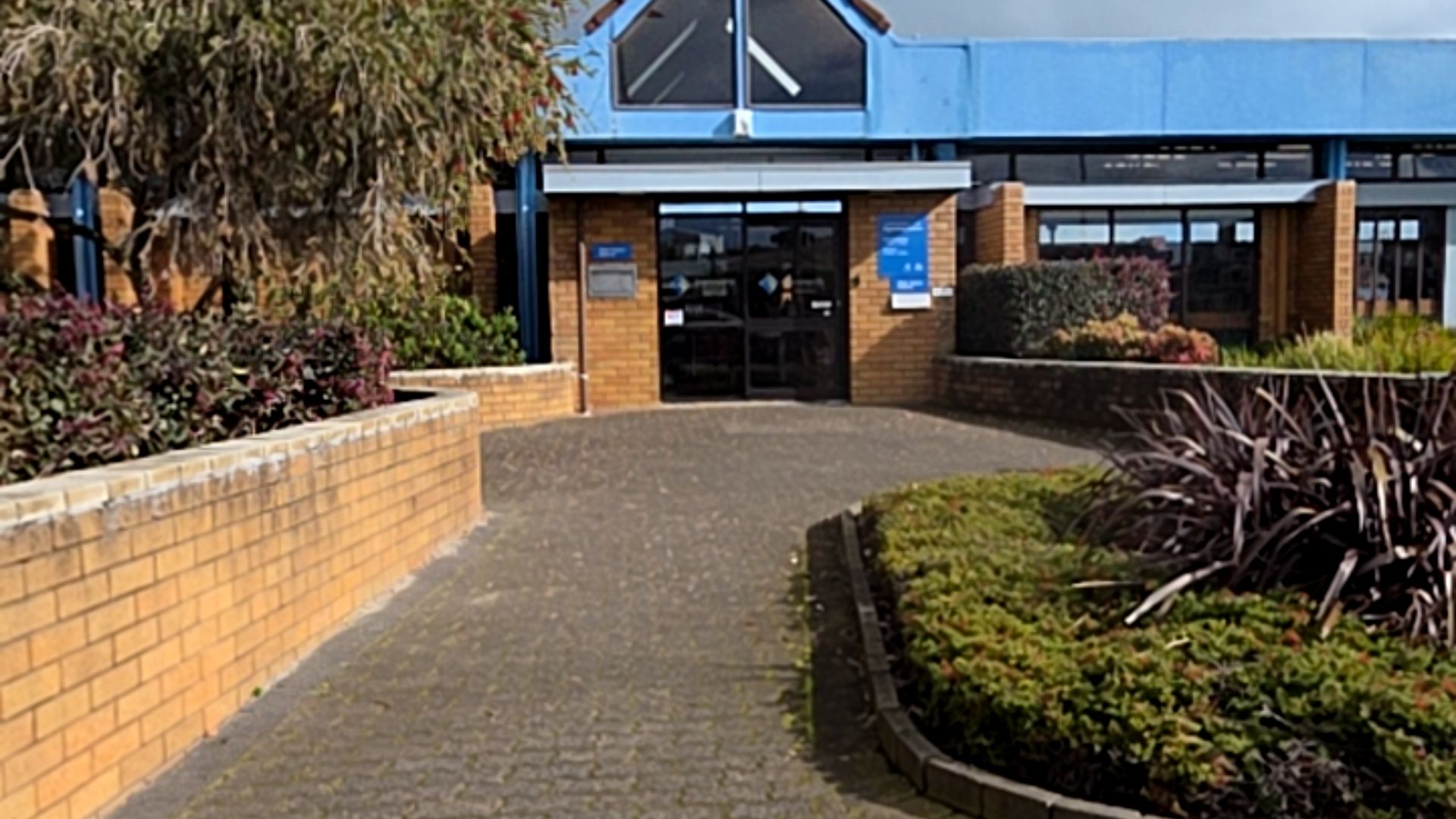 Wide, level brick-paved pathway leads to glass entrance doors with overhead sensors. The building features light brown brick walls and blue signage. No steps visible on the flat approach.