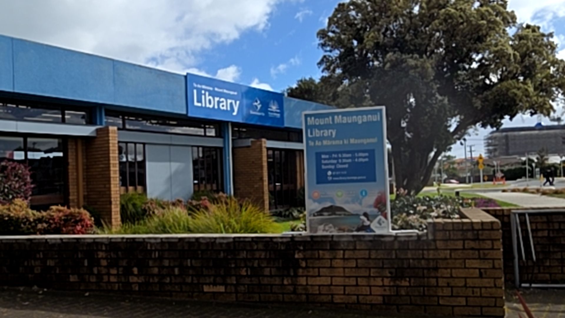 Blue building with 'Library' sign and a large white information board on a low brick wall. Level paved area leads towards glass entrance doors set behind brick pillars.