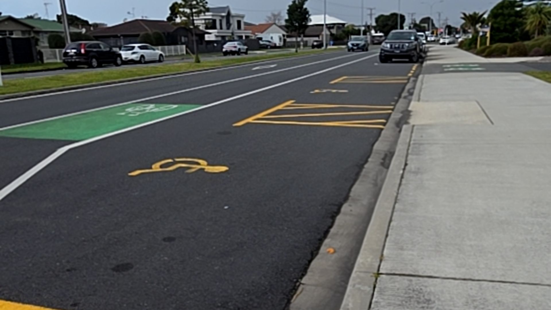 Accessible parking bay with yellow wheelchair symbol and hatched zone. A concrete kerb ramp leads to a level footpath. A green cycle lane sits adjacent to the asphalt road parking space.