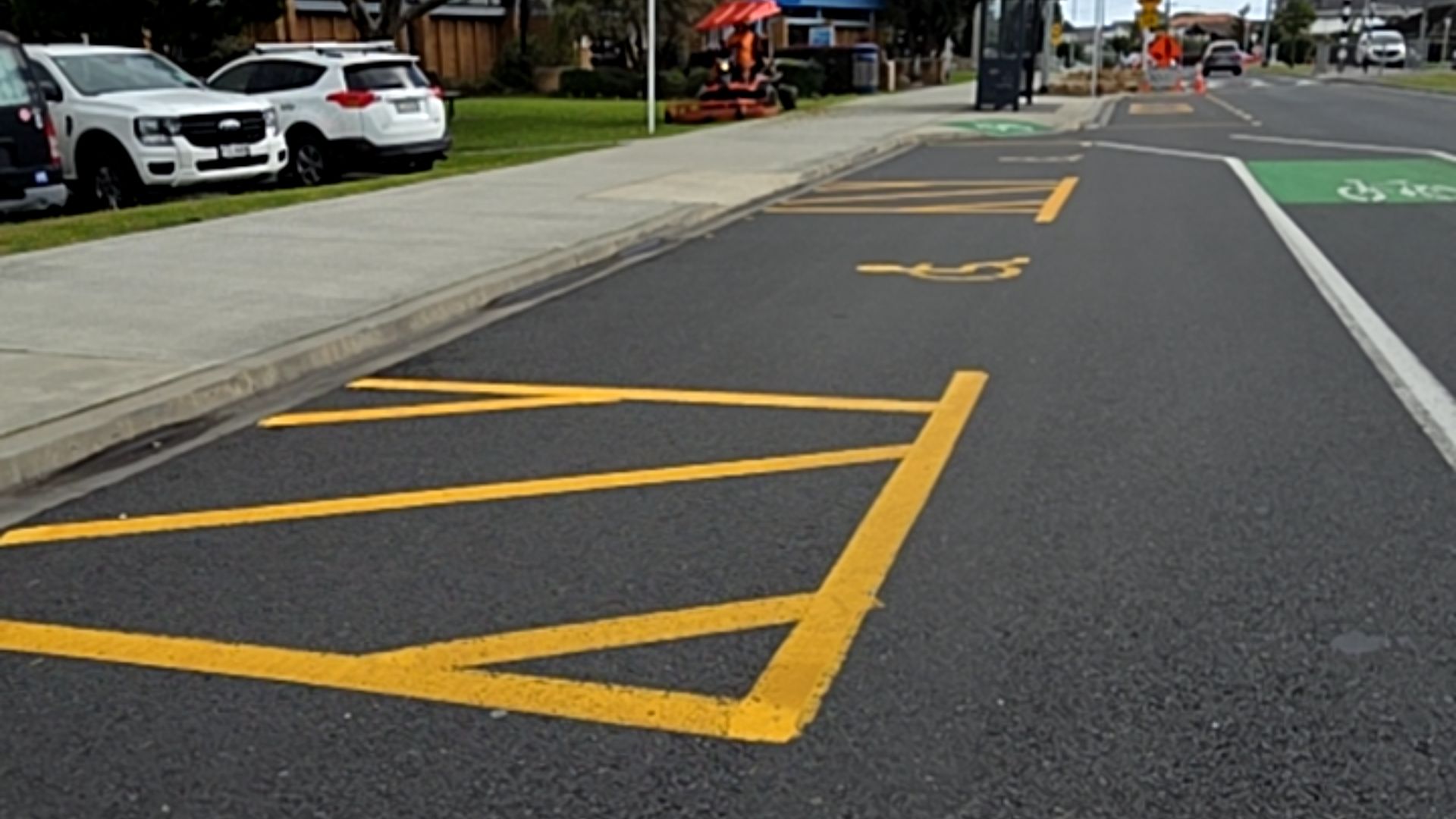 On-street accessible parking with yellow wheelchair symbols and hatched loading zones on level asphalt. Parallel wide concrete footpath features a low kerb for access.